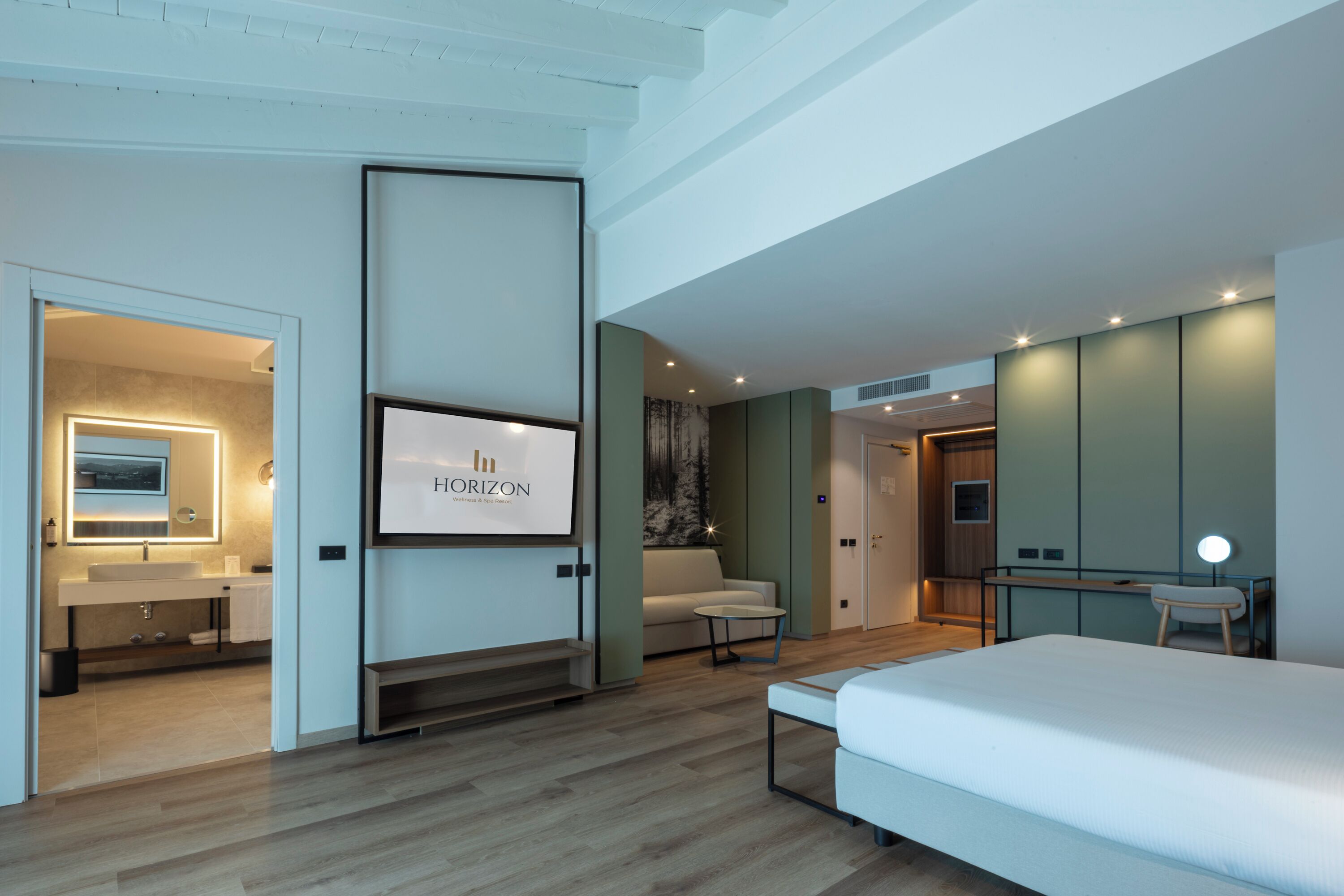 A modern bedroom with a large bed, a desk with a chair, a sofa, and a bathroom with a mirror and sink.