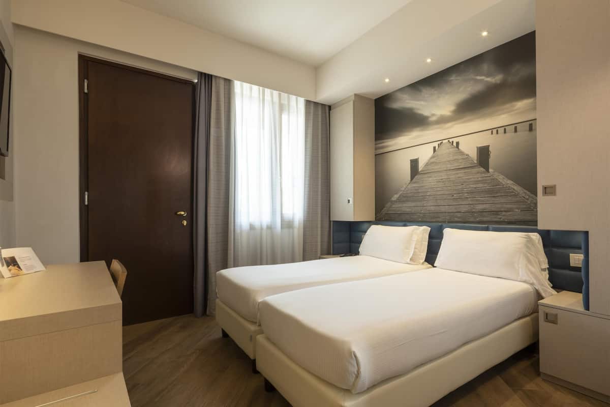 A hotel room with two beds, a wooden floor, a large window with curtains, a painting of a bridge, and a desk with a chair.