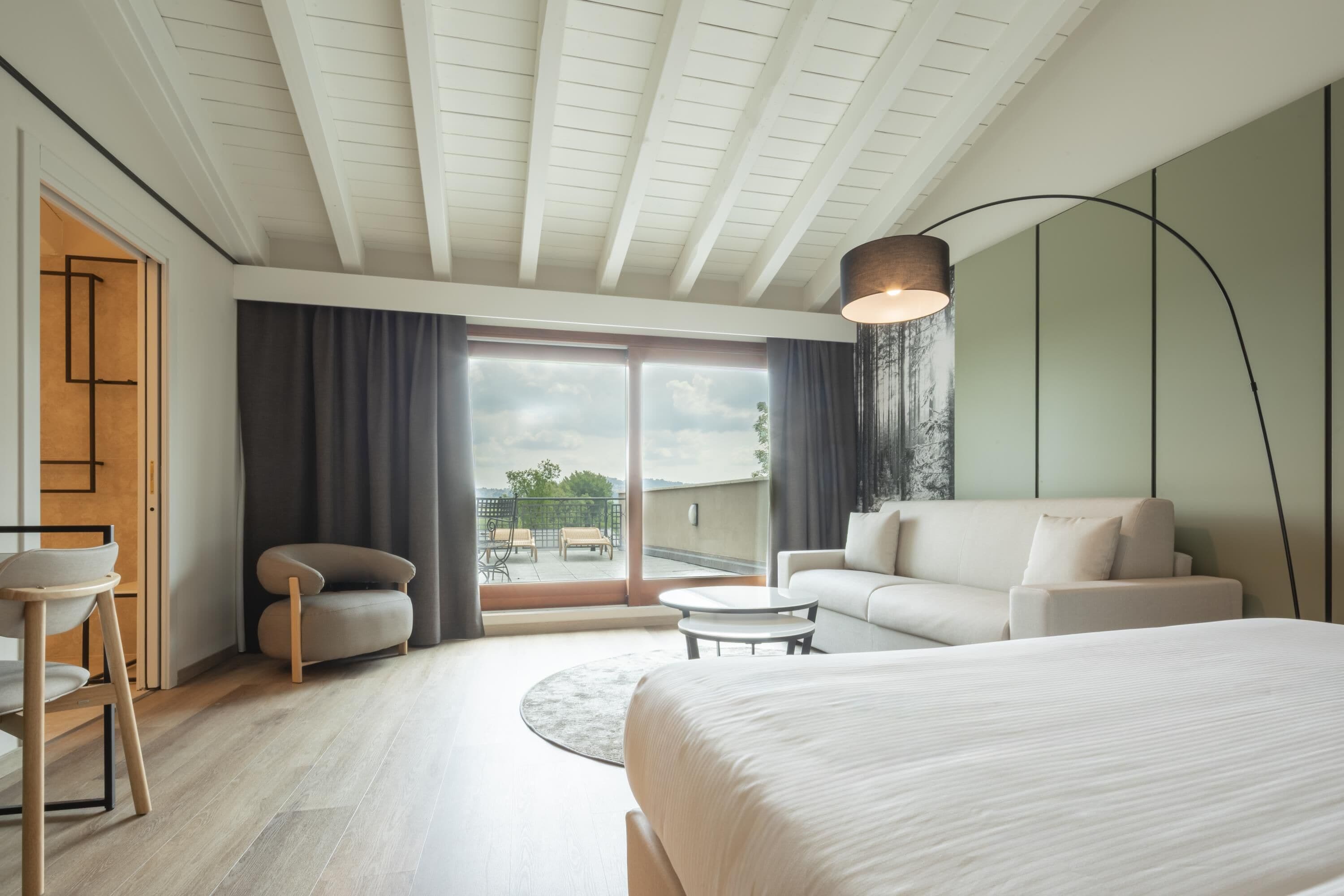 A modern hotel room with a large bed, a sofa, a small table, and a view of the outdoors through a sliding door.