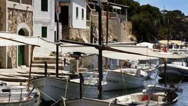 A marina with boats docked, a white building with a balcony, and a stone wall. - Hotel Rocamar (Santanyi)