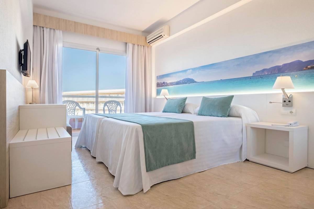 A hotel room with a large bed, a seating area, a TV, and a painting of a coastal scene.