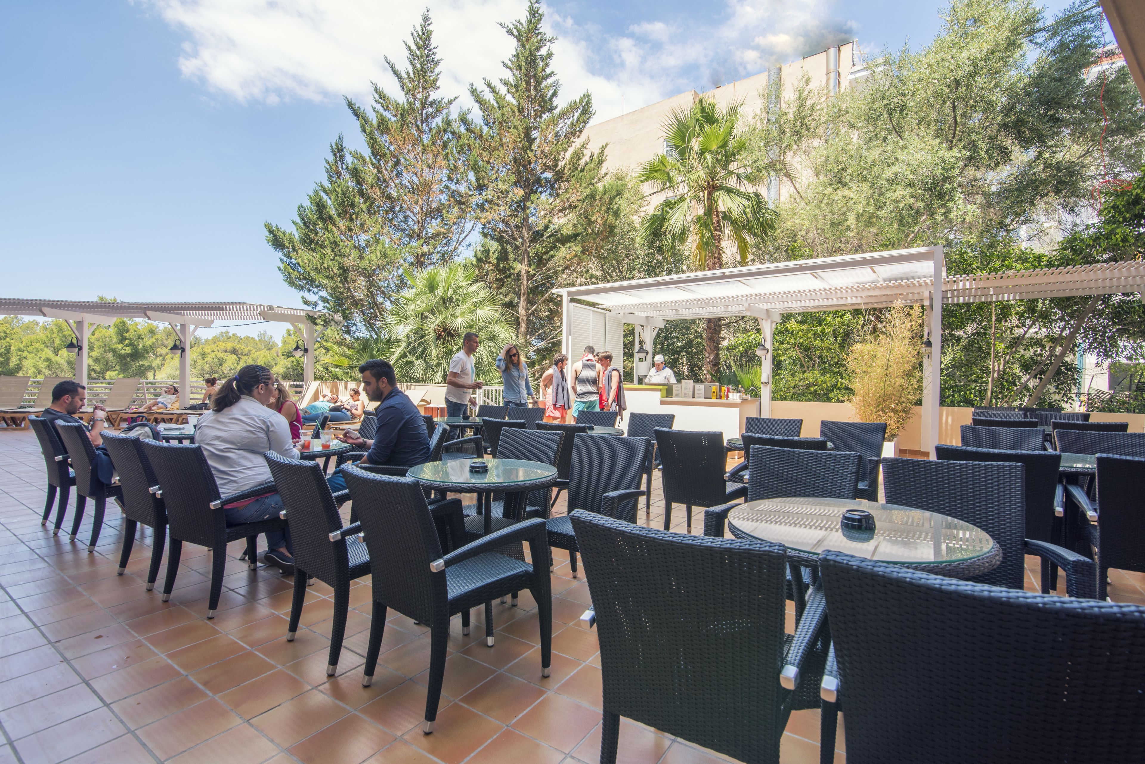 Un patio al aire libre con mesas y sillas redondas, rodeado de árboles y un edificio al fondo.