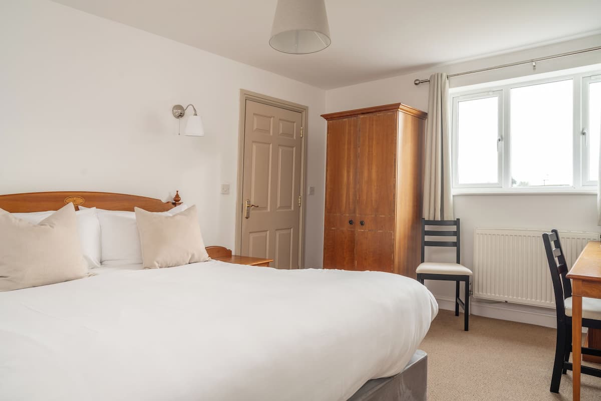 A bedroom with a large bed, a wooden wardrobe, a chair, a table, and a window with curtains.