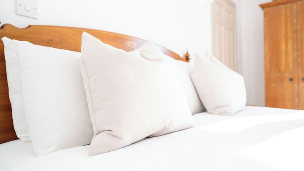 Ein Bett mit weißer Bettwäsche und Kissen, ein Holz-Kopfbrett und eine weiße Wand mit Lichtschalter.