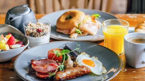 Ein Frühstückstisch mit verschiedenen Speisen, darunter Eier, Würstchen und ein Bagel, sowie ein Glas Orangensaft und eine Tasse Kaffee.