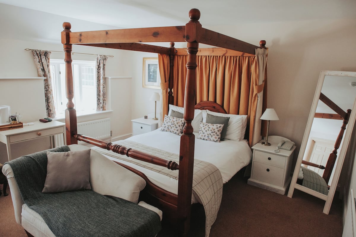 A four-poster bed with a wooden frame, a bedside table with a lamp, and a chair with a cushion.