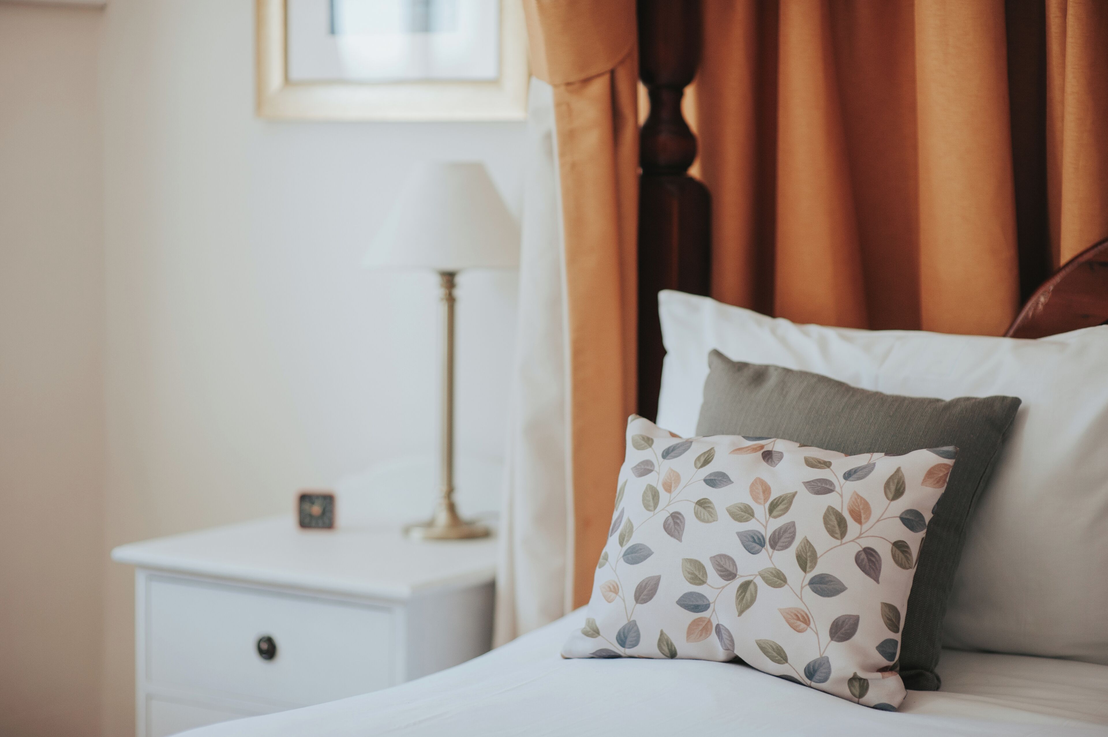 A neatly made bed with a patterned pillow and a plain pillow, a bedside lamp, and a small clock on a white nightstand.