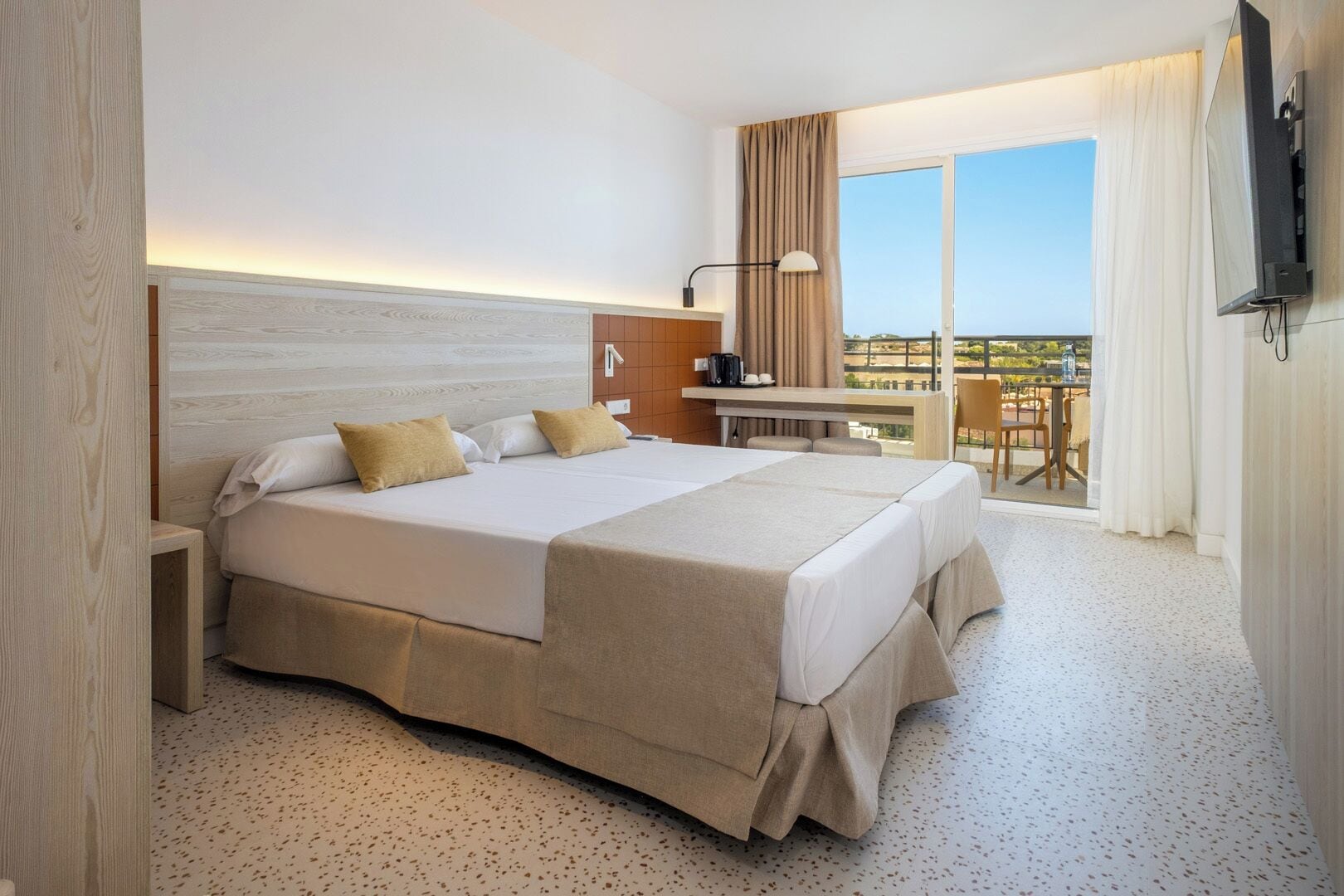 Habitación de hotel con una cama grande, un escritorio con silla, una televisión y un balcón con mesa y sillas.