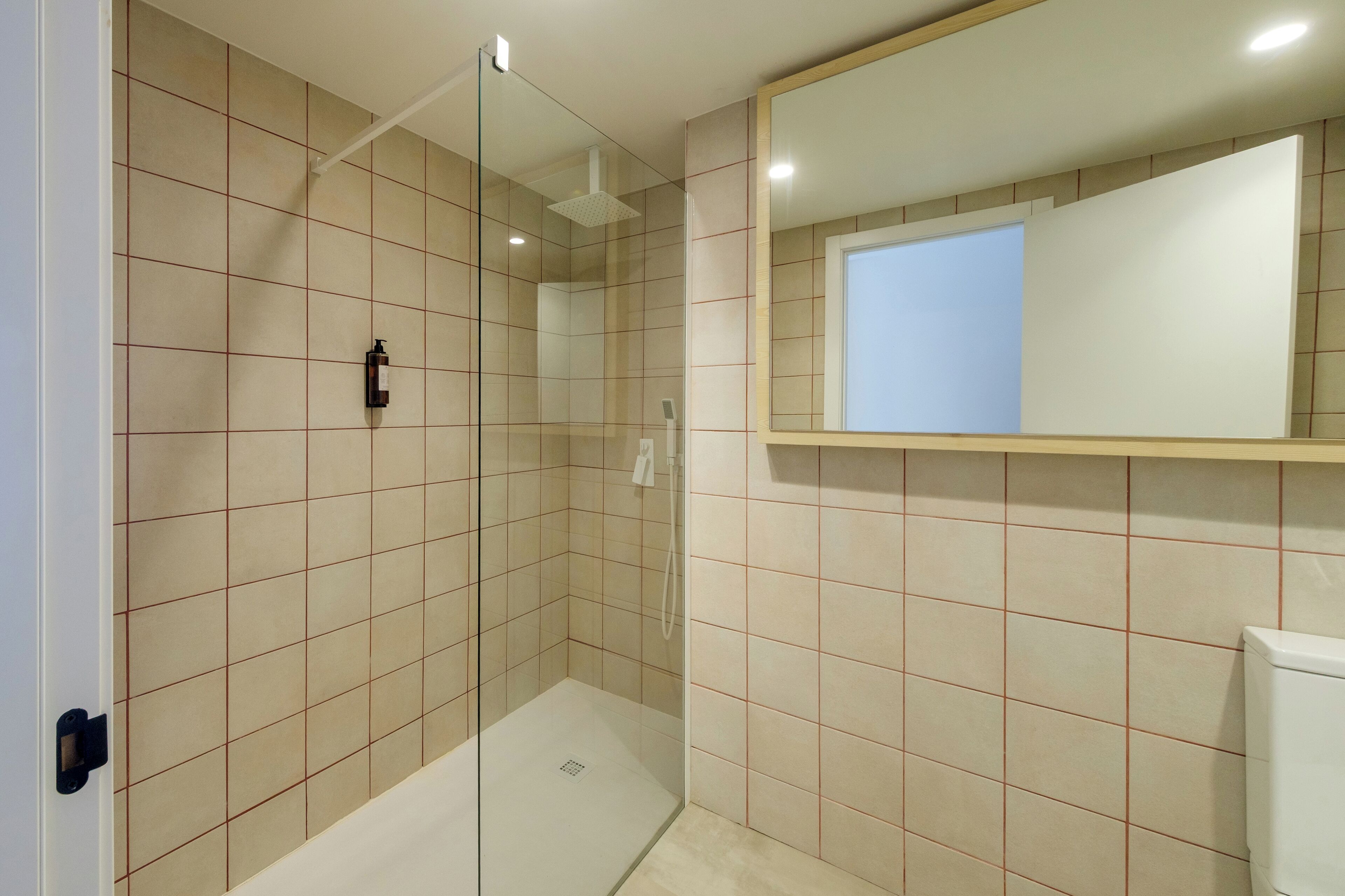 Ein modernes Badezimmer mit beigen Fliesen, einer Duschkabine aus Glas, einer weißen Toilette und einem Spiegel über dem Waschbecken.