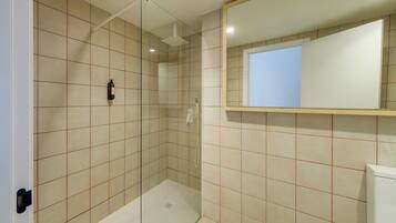Ein modernes Badezimmer mit beigen Fliesen, einer Duschkabine aus Glas, einer weißen Toilette und einem Spiegel über dem Waschbecken.