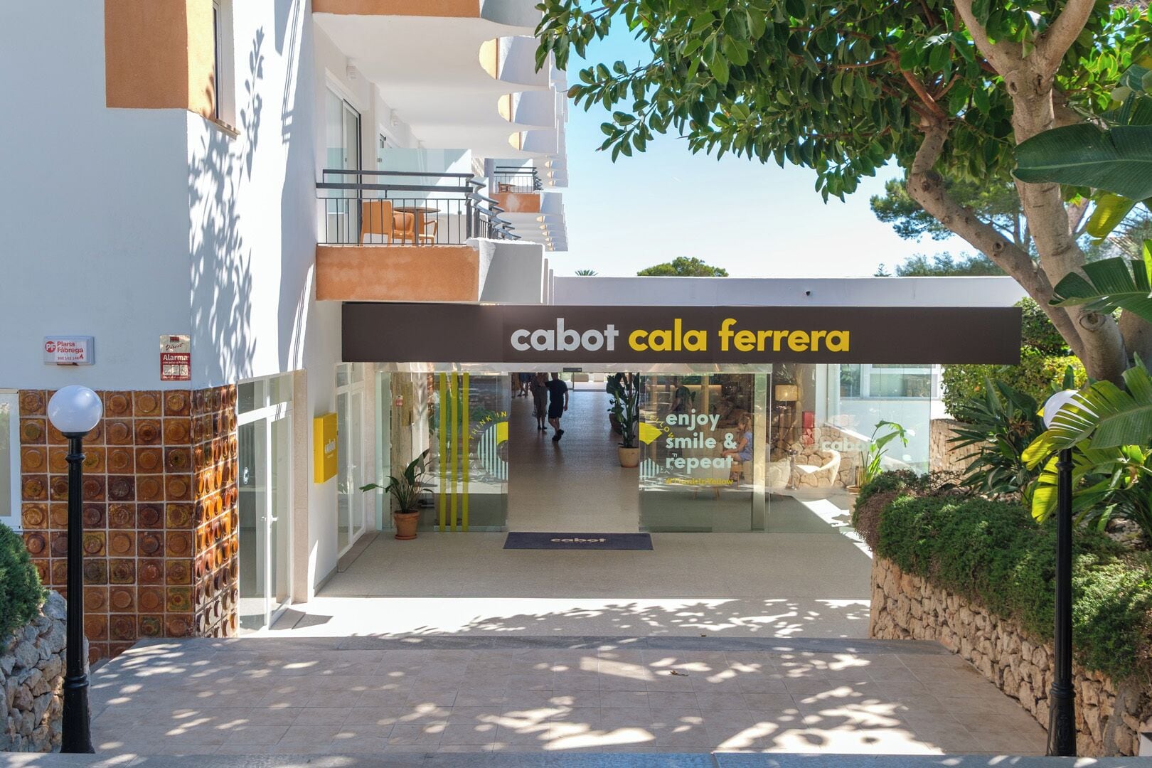 Een gebouw-ingang met een bord waarop 'cabot cala ferrera' staat, een glazen deur en een pad dat naar binnen leidt.
