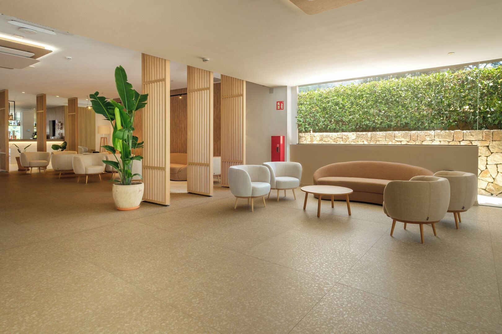 Eine geräumige Lobby mit einer großen Pflanze in einem Topf, einer Sitzecke mit Sofas und Sesseln sowie einer Steinwand mit Bepflanzung.