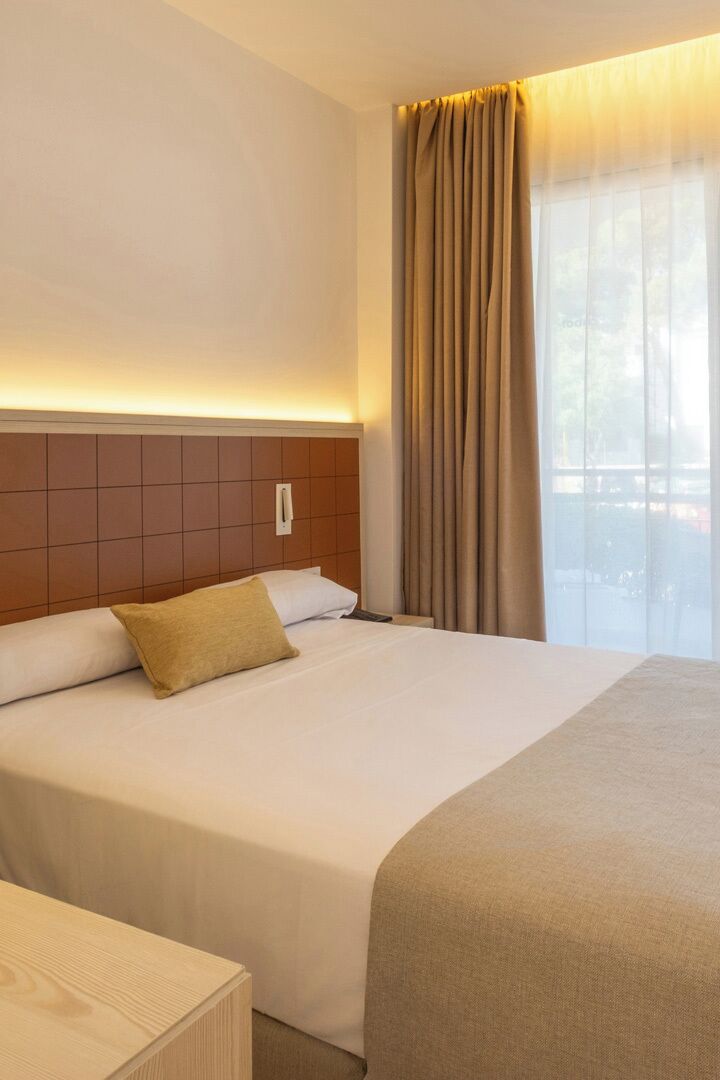 Una habitación de hotel con una cama grande, cabecera, mesita de noche y ventana con cortinas.