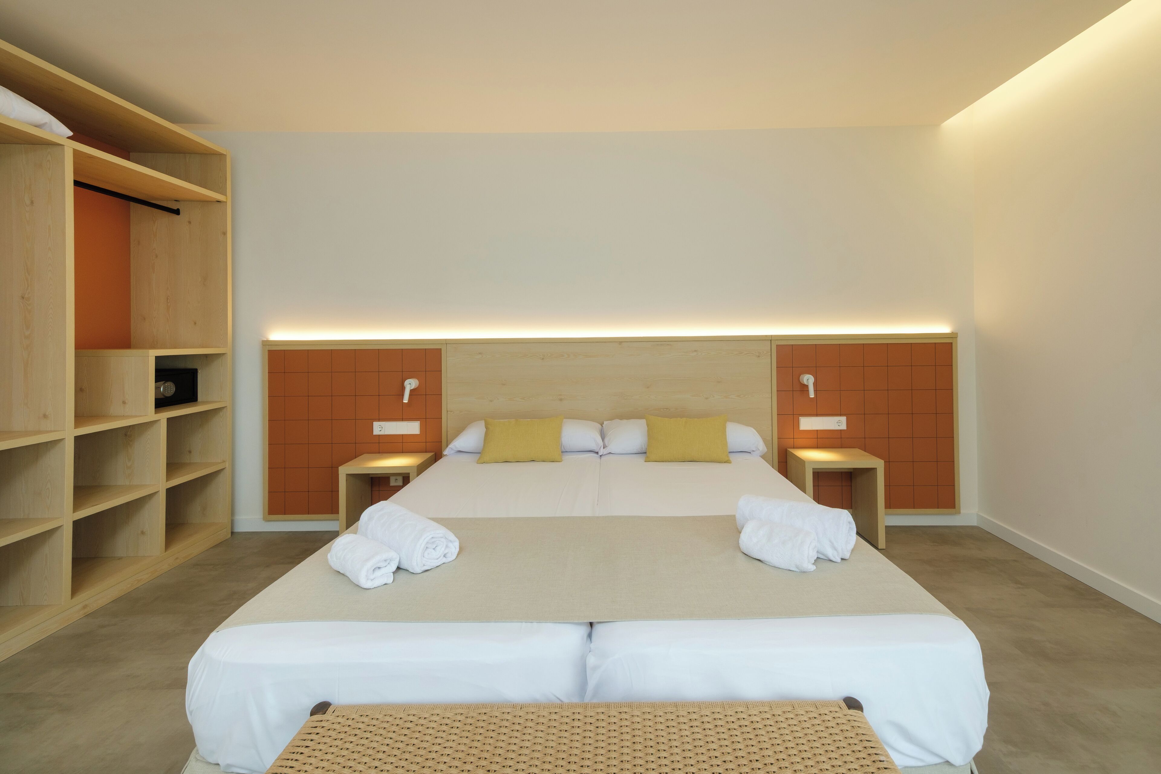 A double bed with white linens, two pillows, and a headboard with a wooden panel design. There are two bedside tables with lamps, and a wooden wardrobe with shelves.