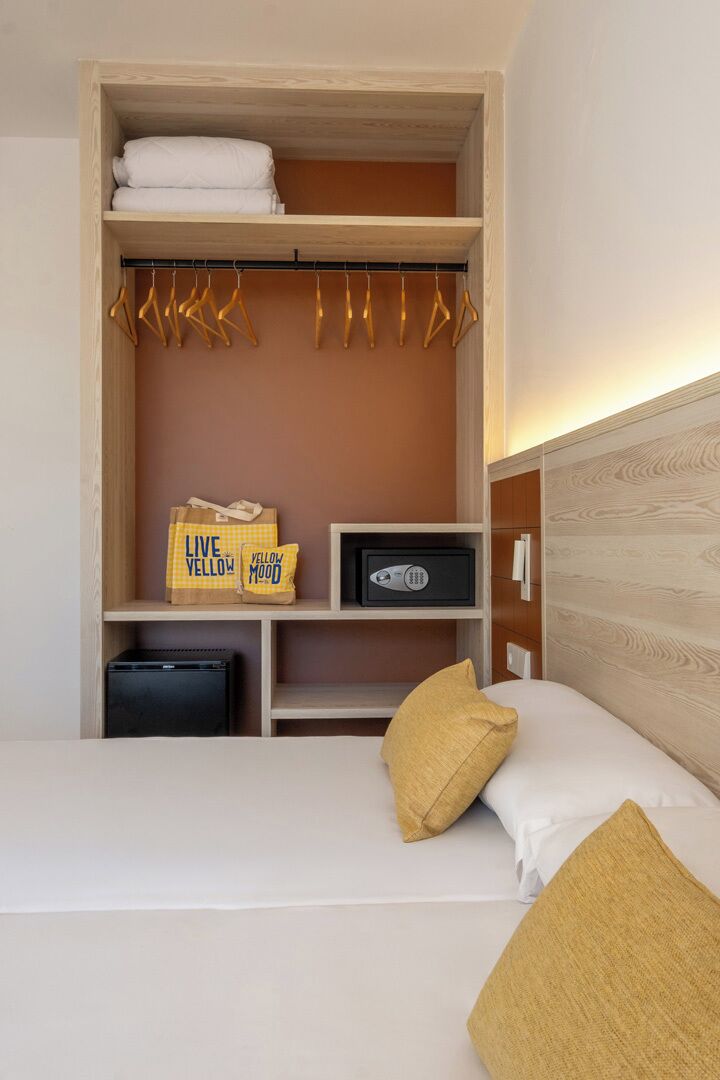 A hotel room with a bed, a wardrobe with hanging clothes, a shelf with a radio, and a small safe.