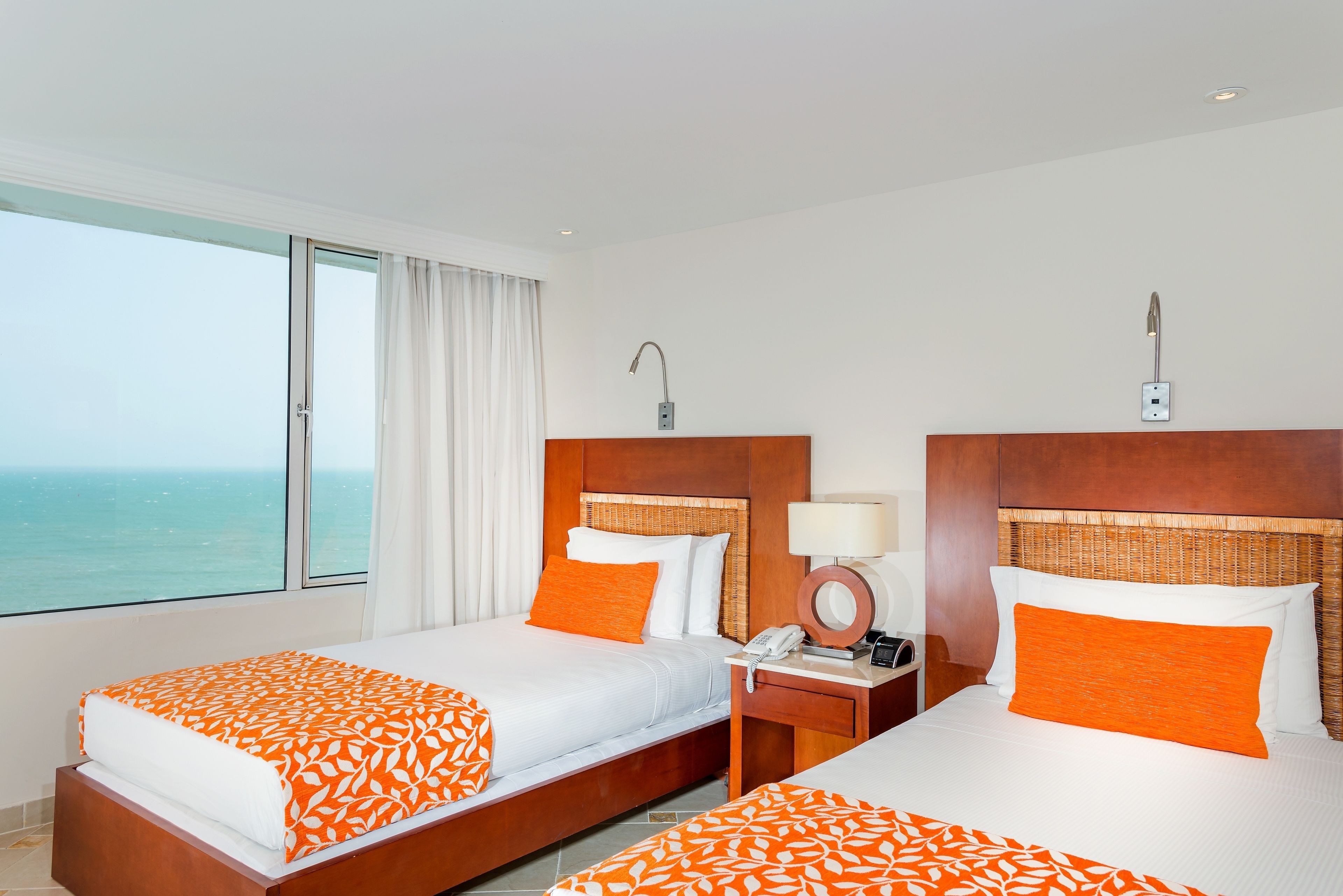 Una habitación de hotel con dos camas, un ventanal con vista al mar, una mesita de noche con una lámpara y un teléfono.