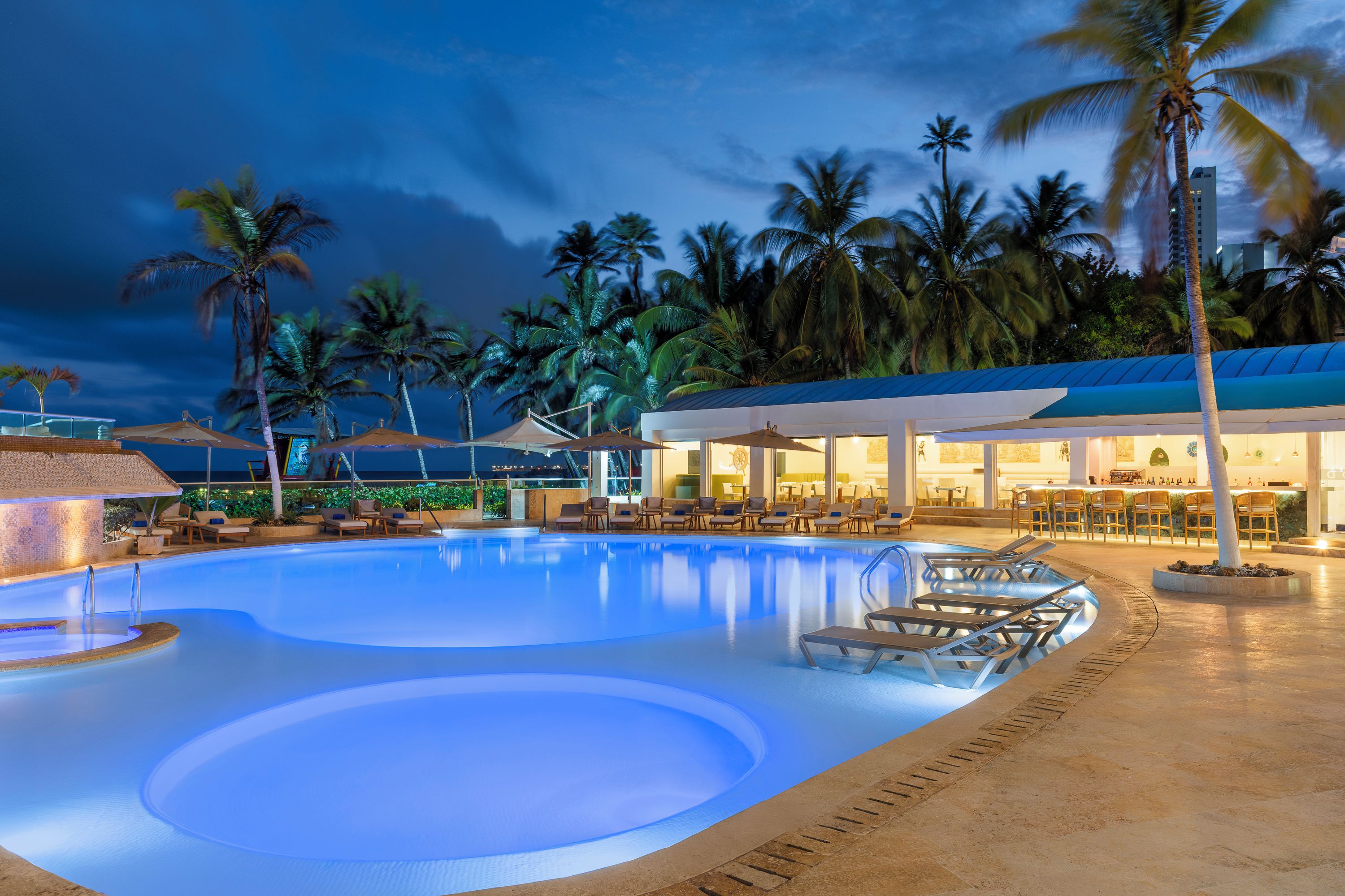 A swimming pool with lounge chairs and umbrellas, surrounded by palm trees and a building with a blue roof.