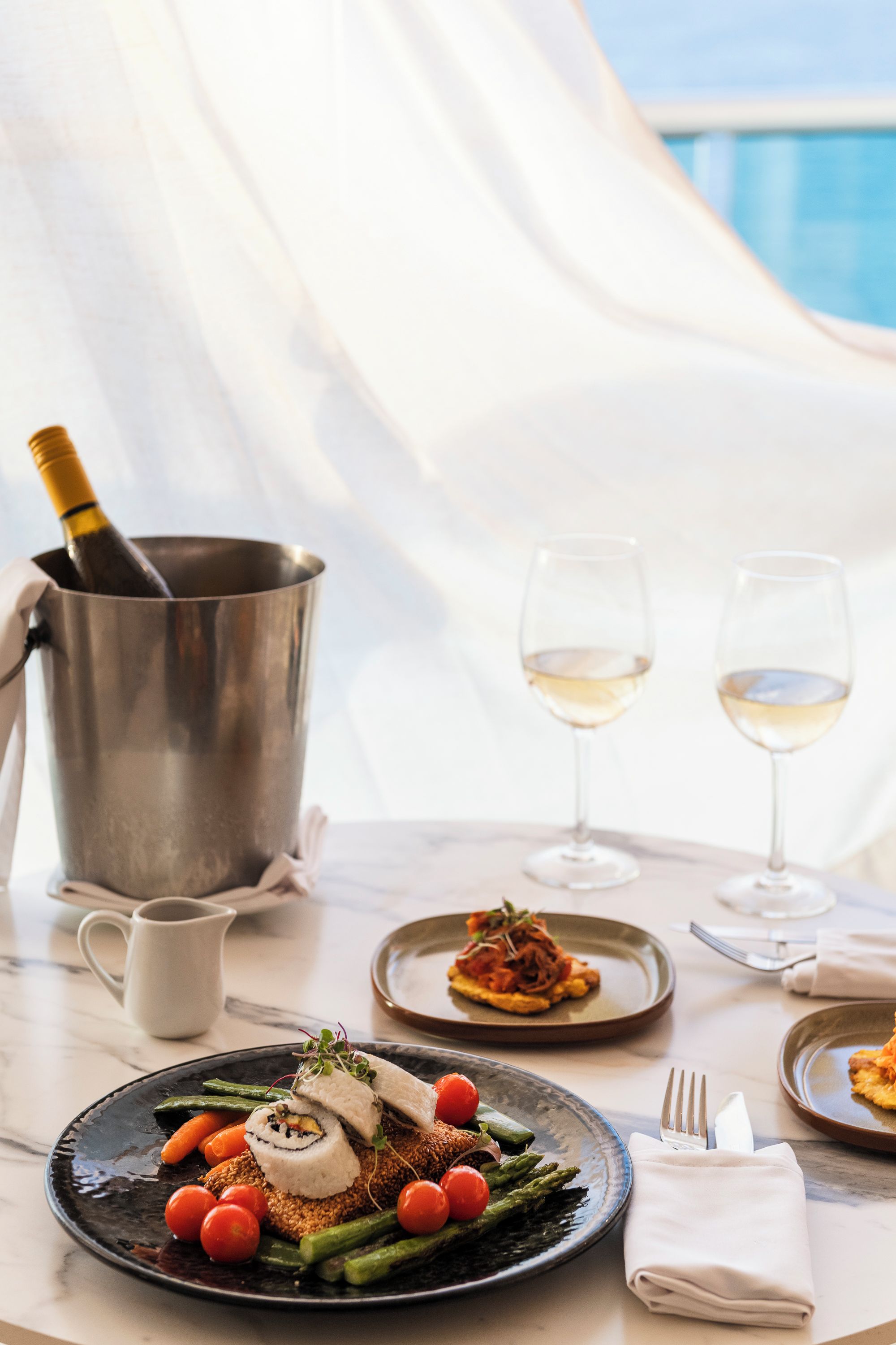 A dining table with a bottle of champagne, two glasses of white wine, and plates of food including a sushi roll and vegetables.