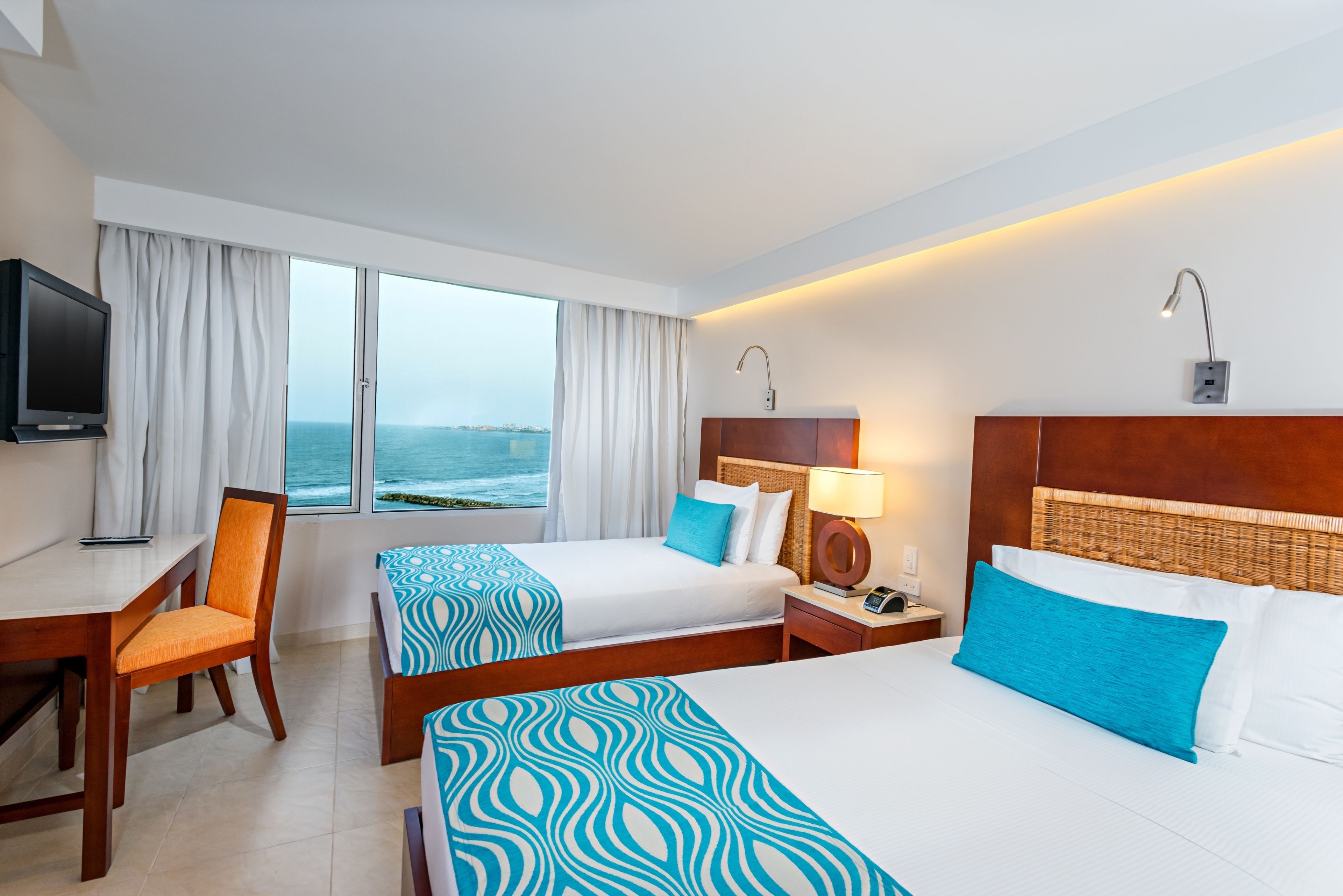 A hotel room with two beds, a desk, a chair, a TV, and a view of the sea.