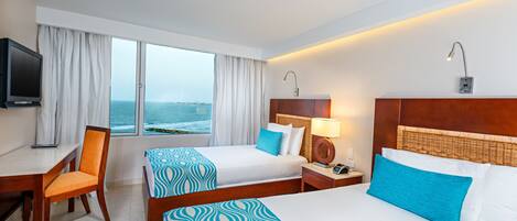 A hotel room with two beds, a desk, a chair, a TV, and a view of the sea.