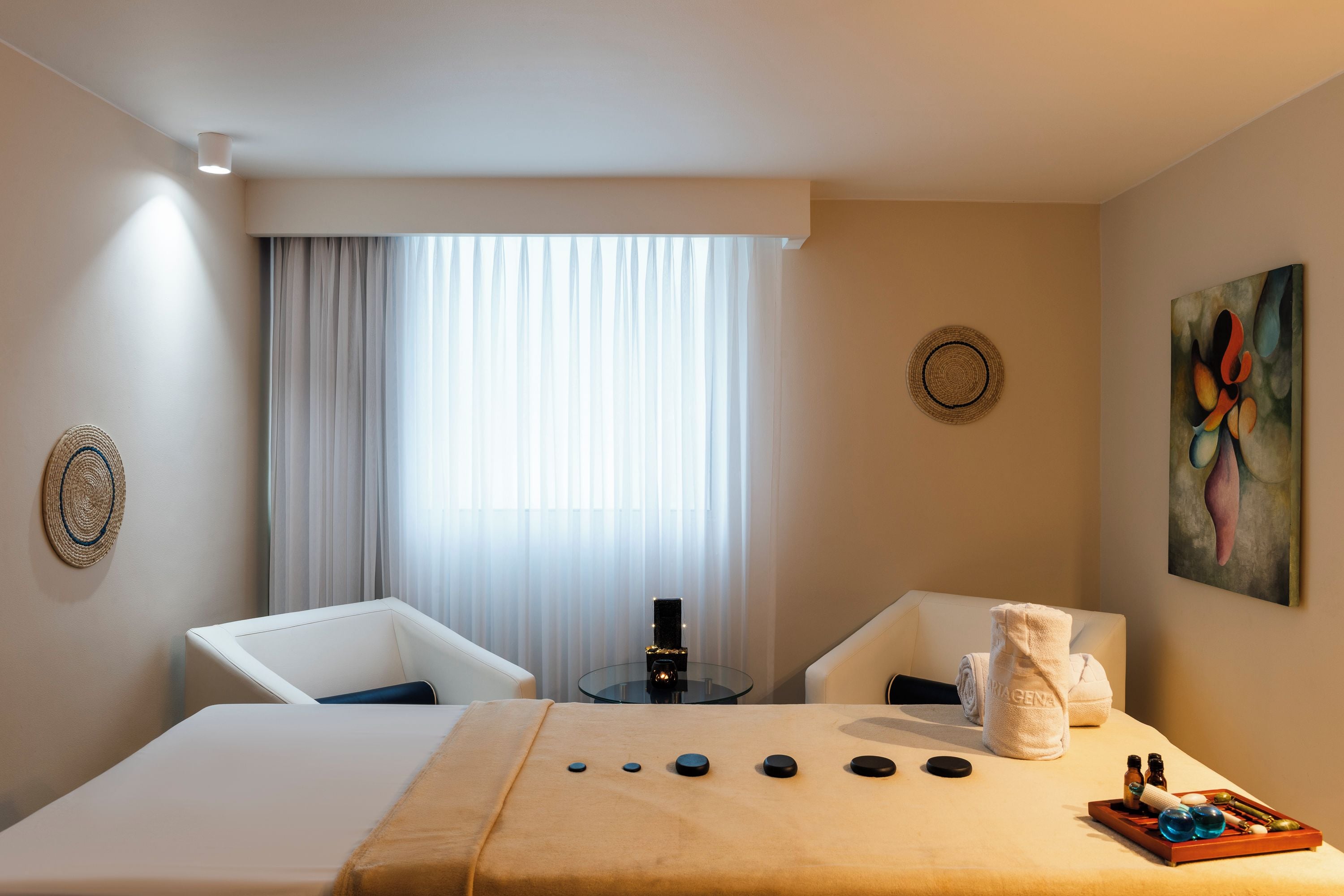 A hotel room with a bed, two chairs, a massage table, a small table with massage tools, and a painting on the wall.