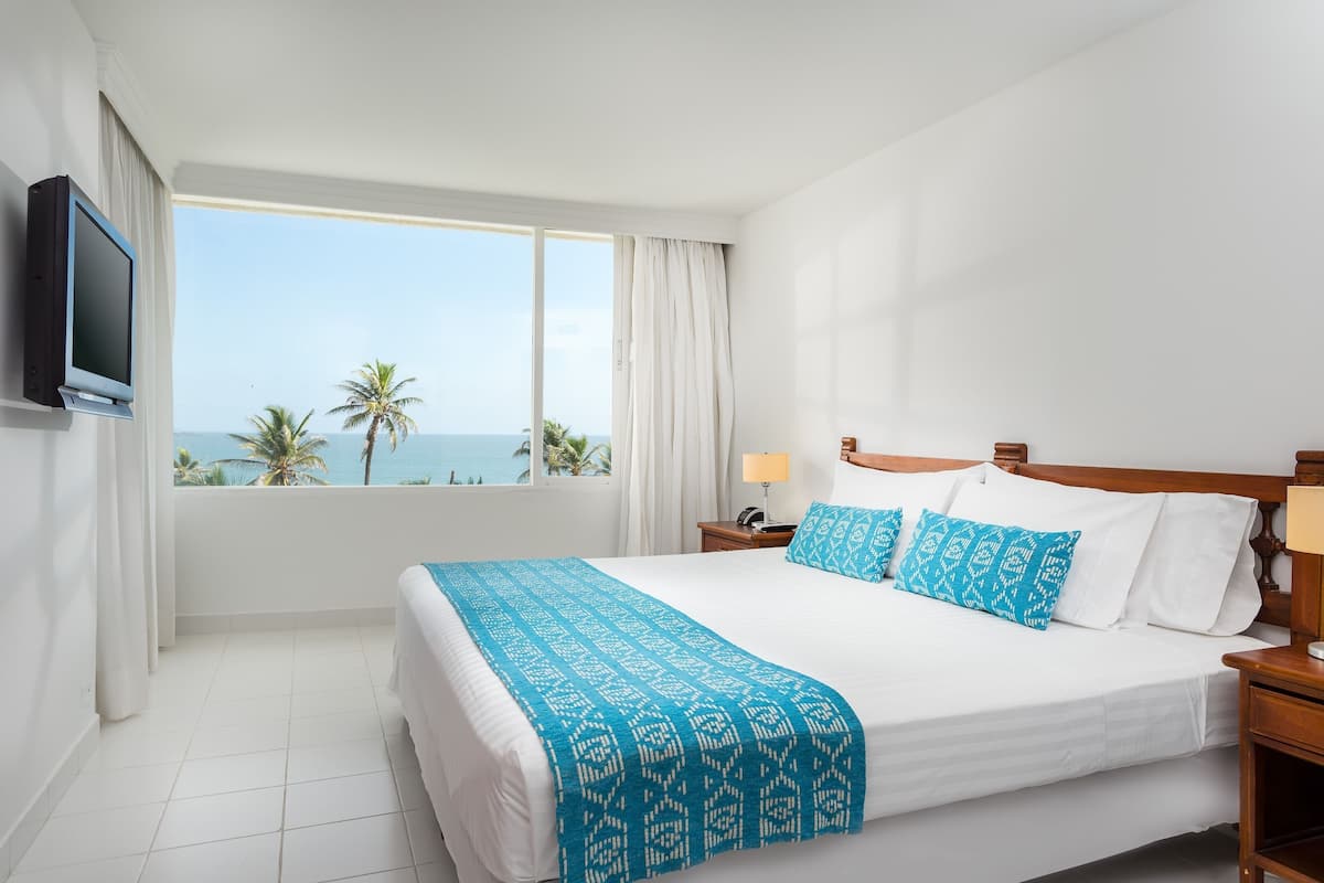 A hotel room with a large bed, a TV mounted on the wall, a nightstand with a lamp, and a view of the ocean through large windows.