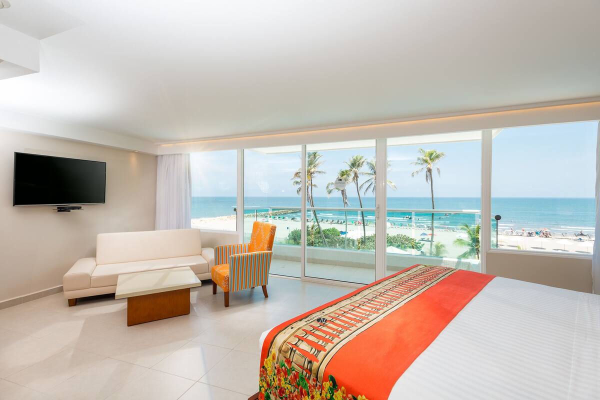 A hotel room with a large bed, a sofa, a chair, a TV, and a balcony with a view of the beach and ocean.