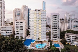 Un hotel de gran altura con múltiples balcones, una piscina en la azotea y palmeras circundantes.