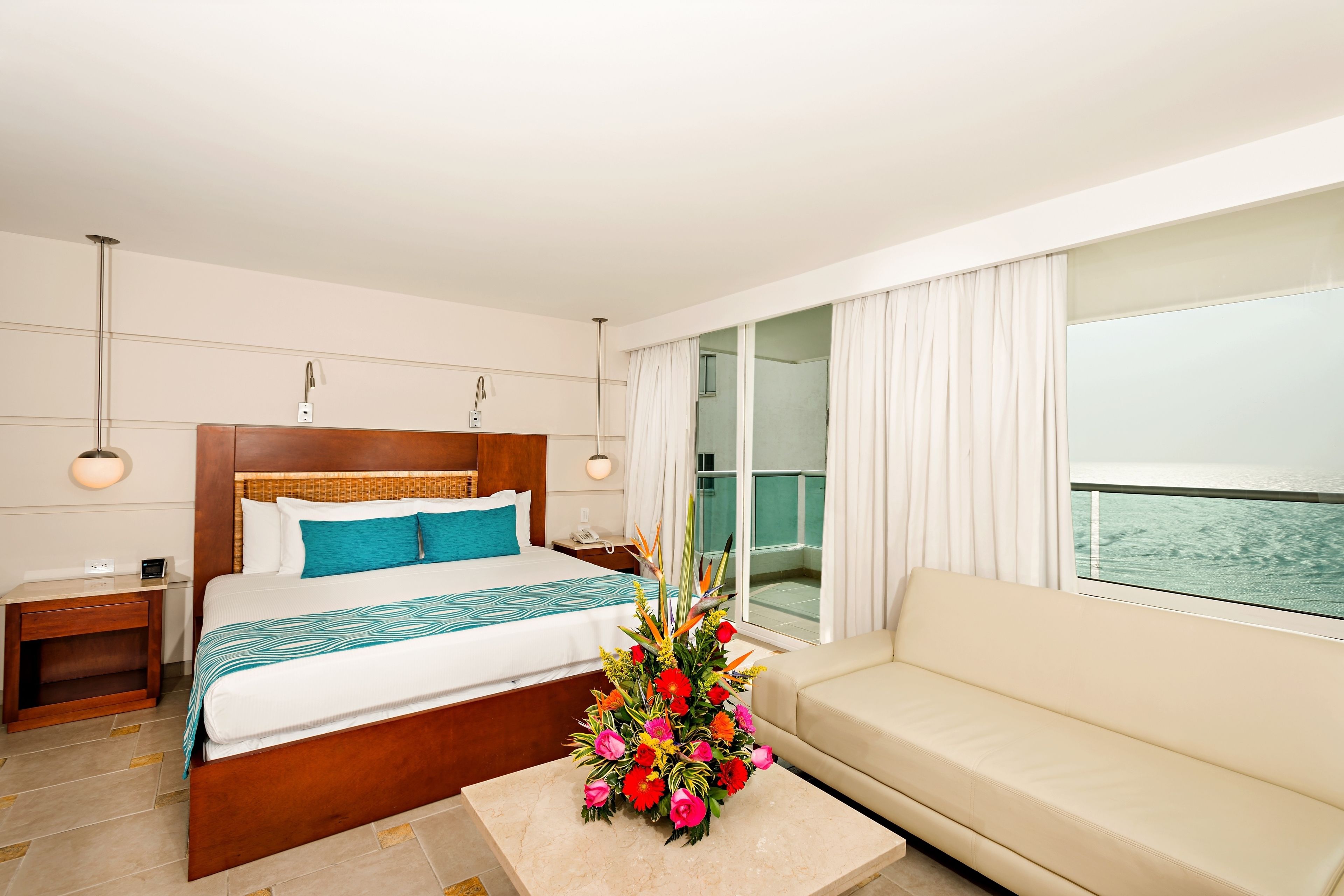 A hotel room with a bed, a sofa, a nightstand, a bedside table, a flower vase, and a window with a view of the sea.