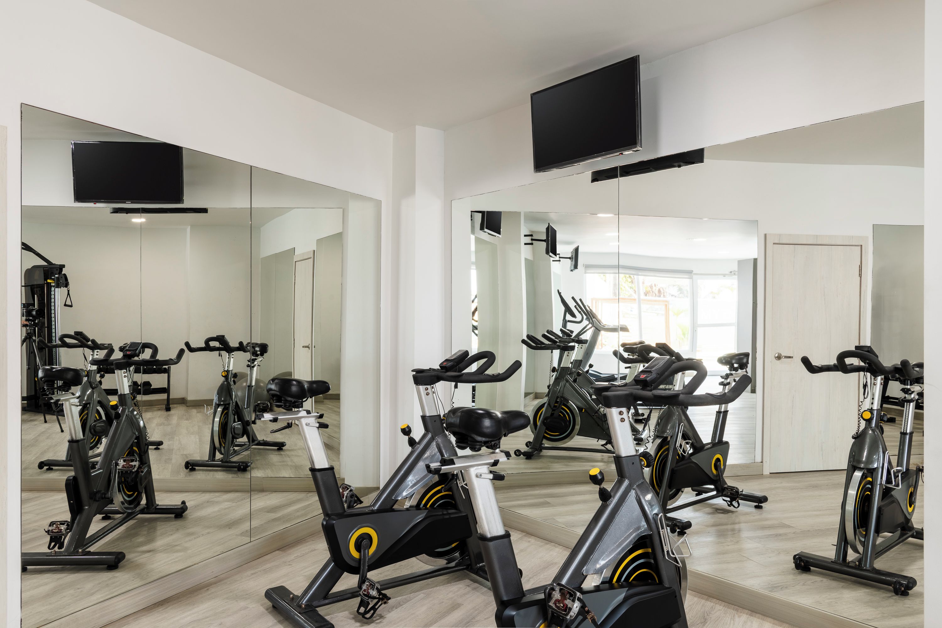A gym with multiple stationary bikes, mirrors, and a TV mounted on the wall.