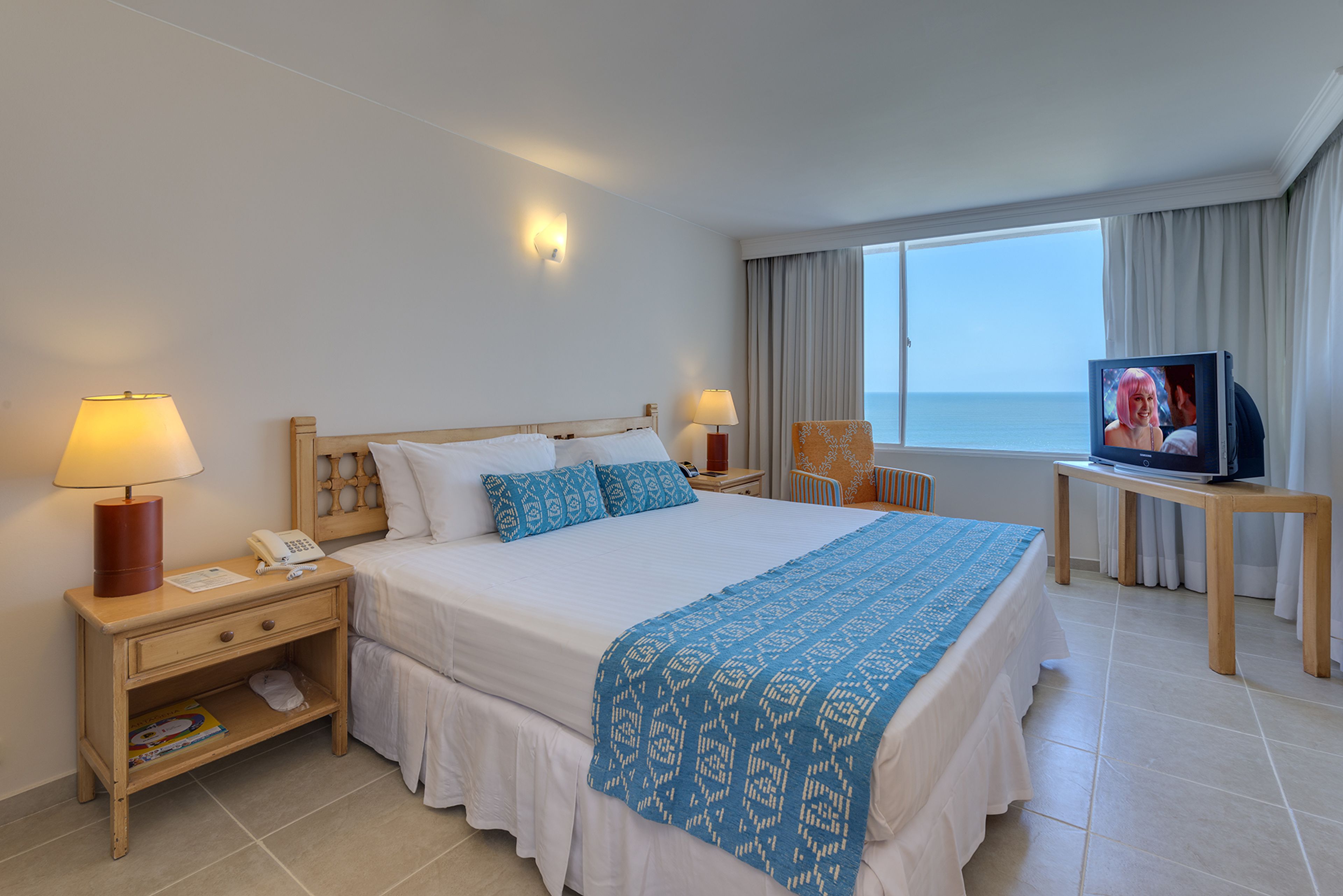 Une chambre d’hôtel avec un grand lit, deux lampes de chevet, une télévision et une vue sur la mer.