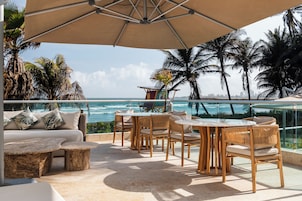 Eine Terrasse mit Meerblick, ausgestattet mit Korbstühlen und einer Couch, und ein großer Sonnenschirm spendet Schatten.