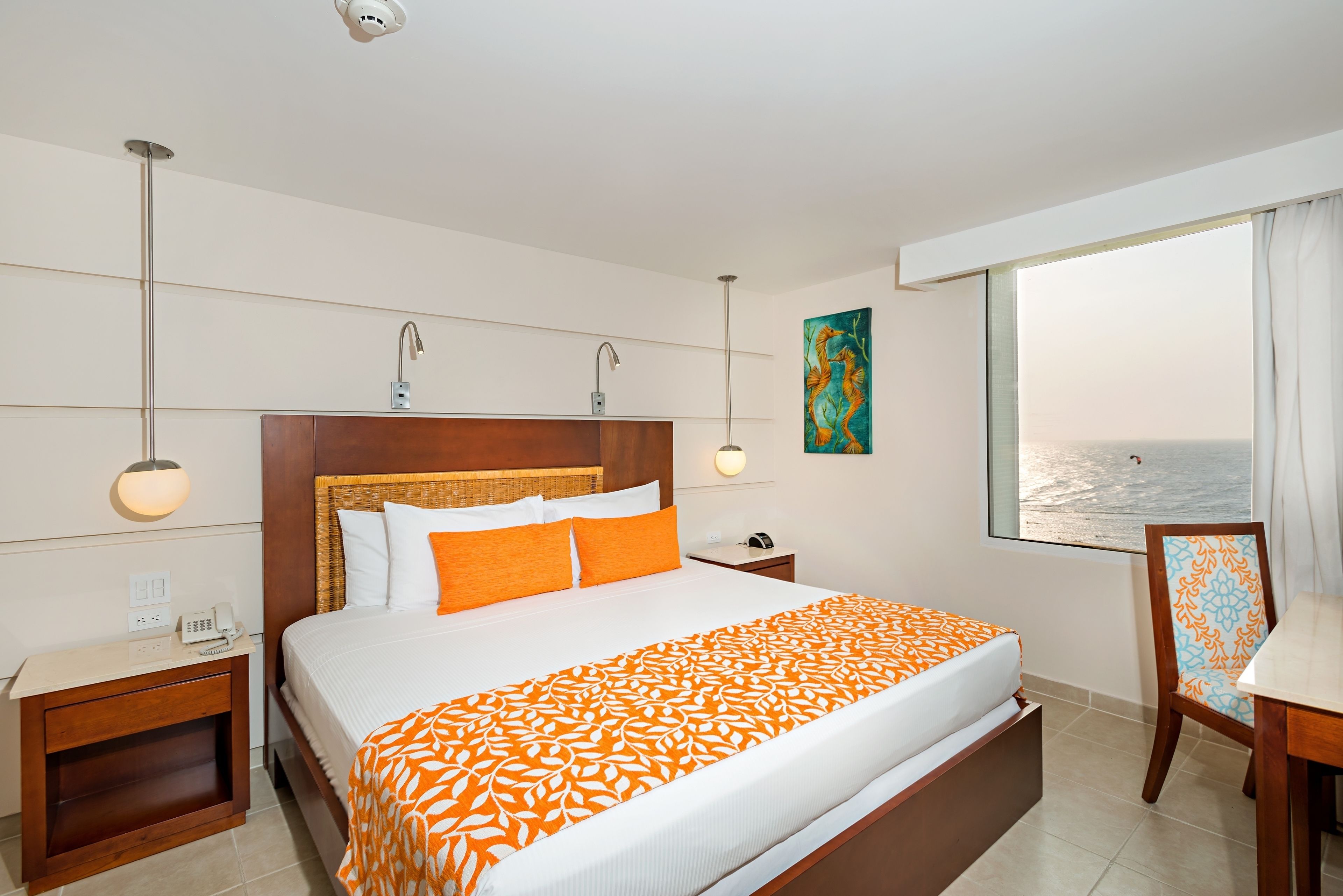 Habitación de hotel con una cama grande, una mesita de noche, un escritorio, una silla, una ventana con vistas al mar y un cuadro en la pared.