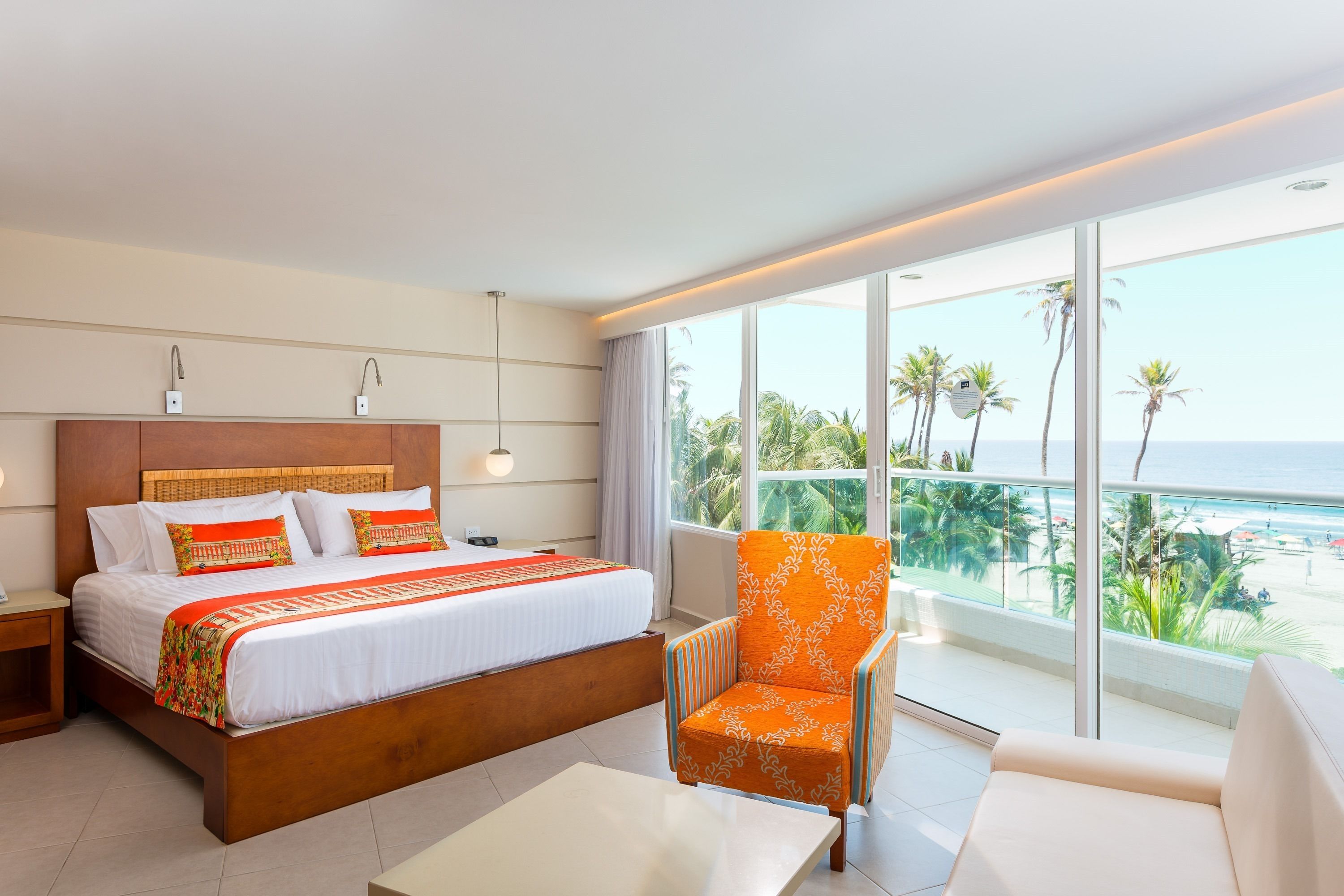 Habitación de hotel con una cama grande, un balcón con vistas a la playa y una silla con un cojín estampado.