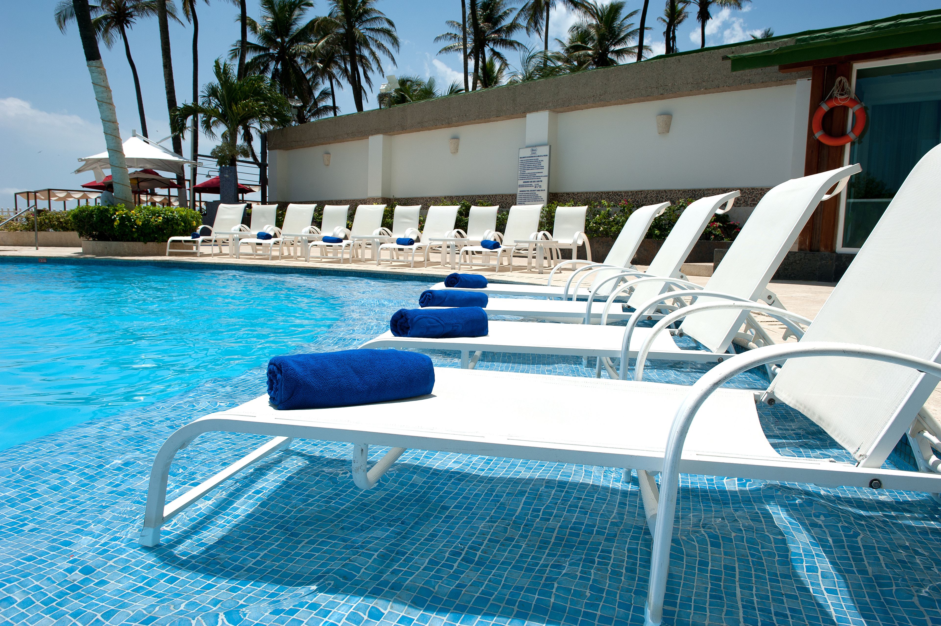 Zona junto a la piscina con sillas de descanso blancas y toallas azules, un flotador salvavidas y palmeras al fondo.
