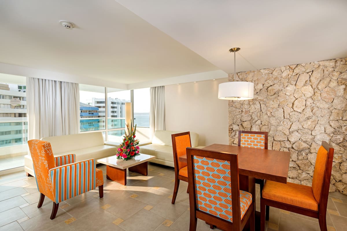A modern hotel room with a stone accent wall, a dining area with wooden furniture, and a large window offering a view of the city.