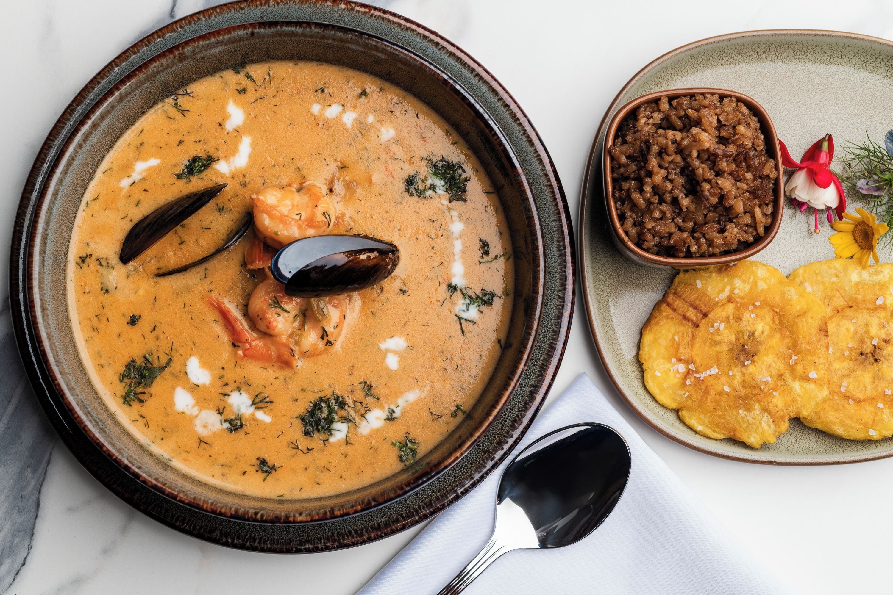 A bowl of seafood soup with mussels and shrimp, a bowl of rice, and a plate of fried plantains.