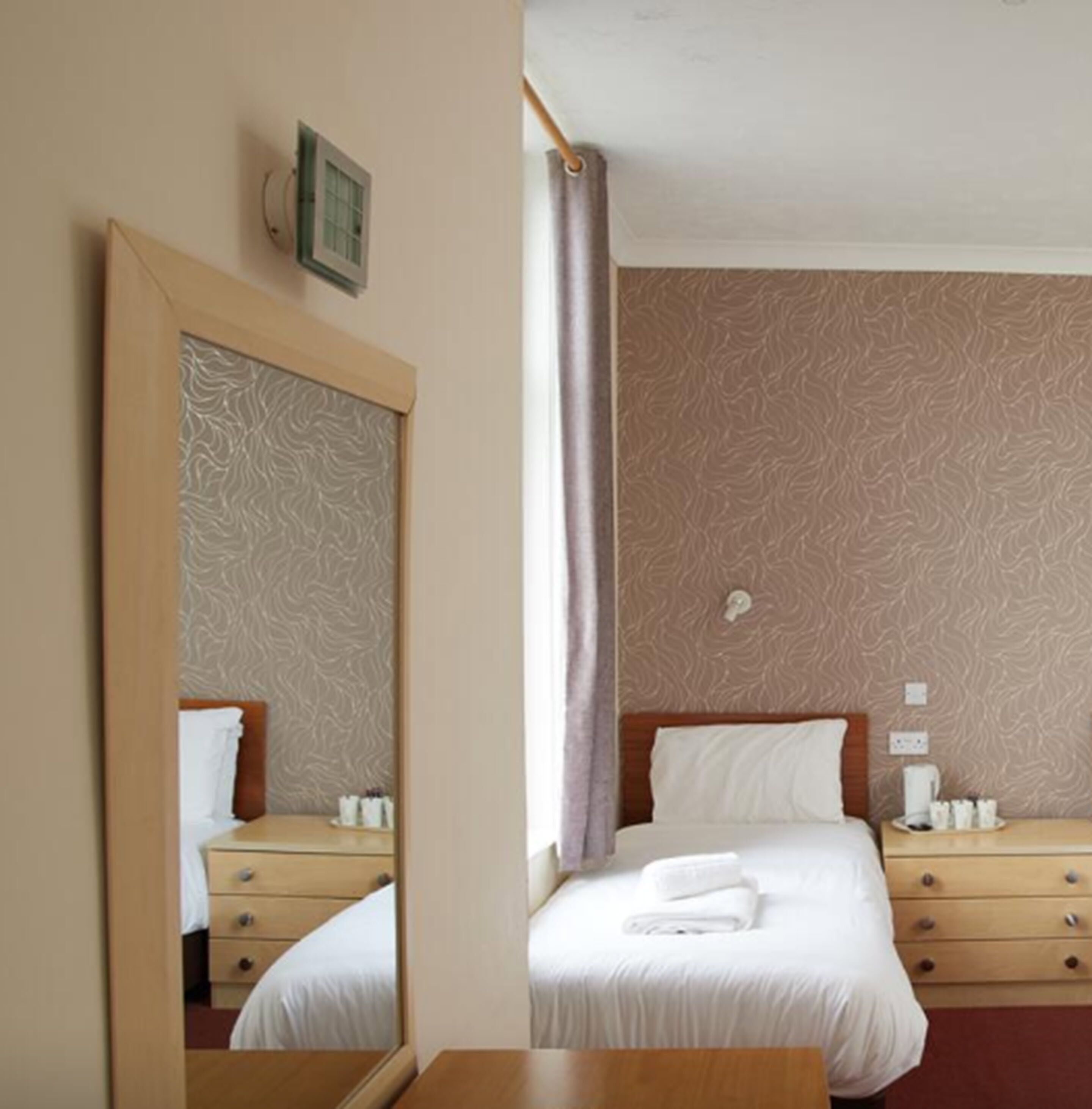Une chambre d’hôtel avec un lit, une commode en bois, un miroir et une fenêtre avec un rideau.