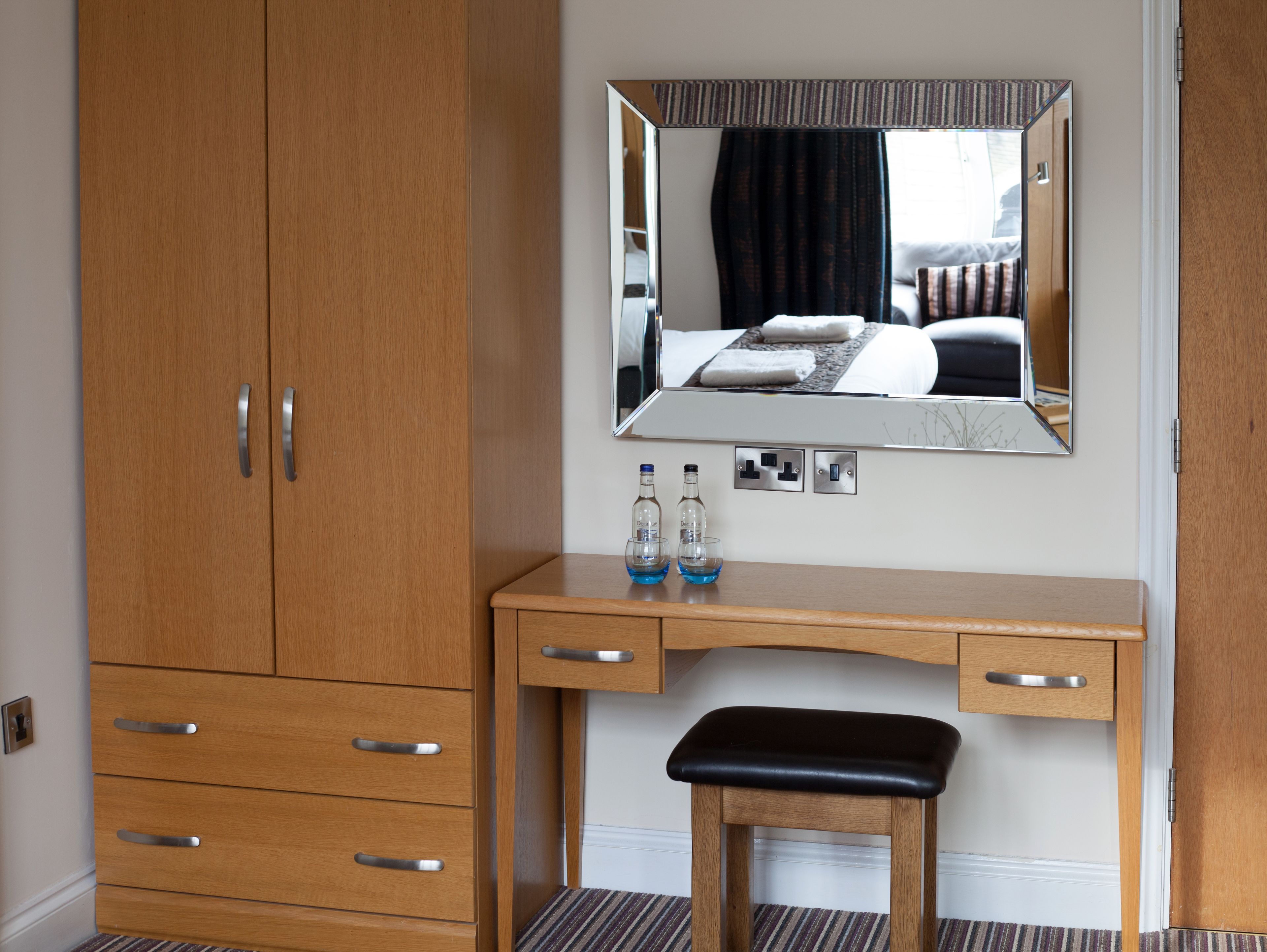 A wooden dresser with drawers and a mirror above it. There is a small stool in front of the dresser.