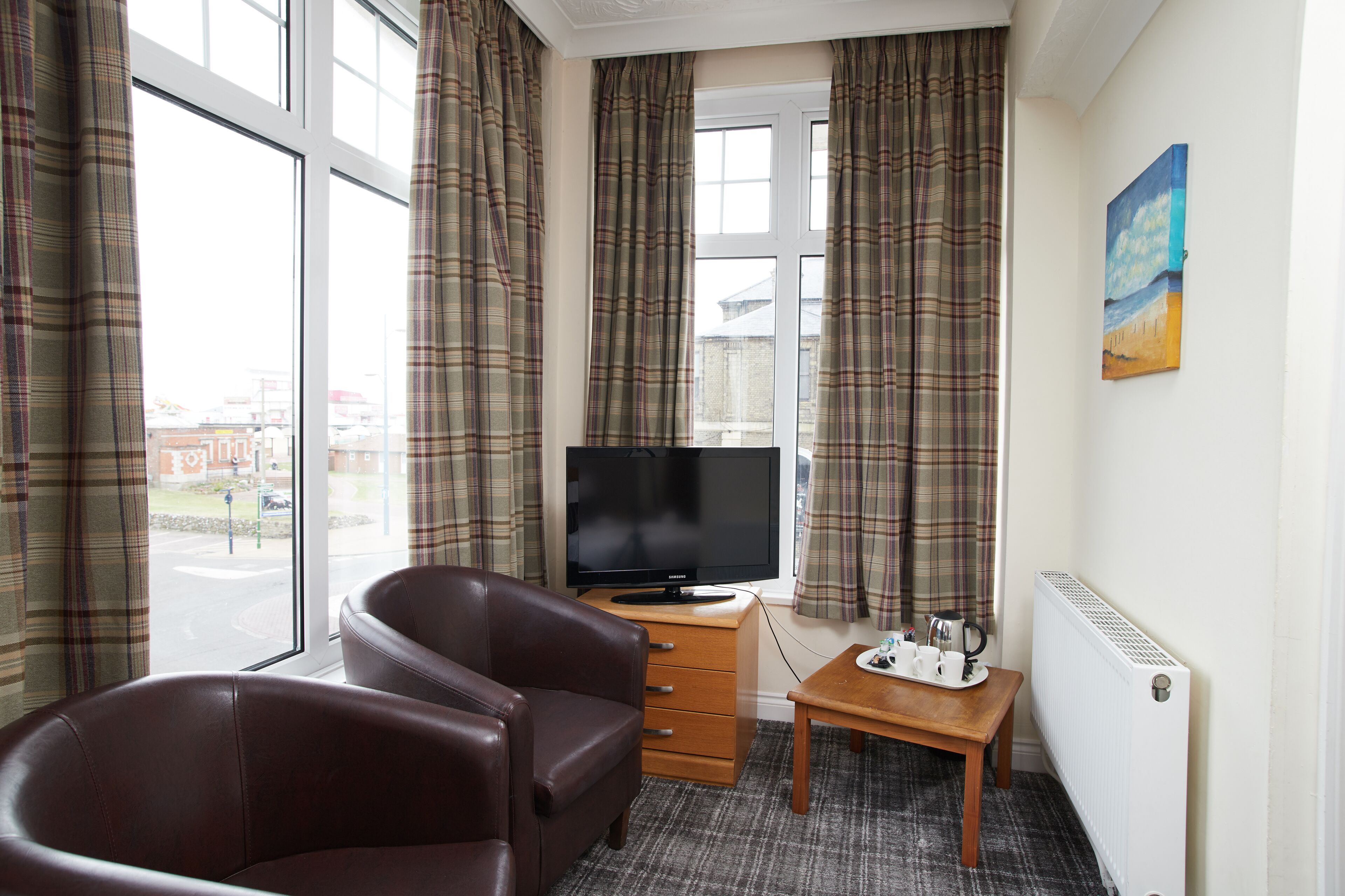 Quarto de hotel com sofá de couro marrom, mesa lateral de madeira com uma bandeja de xícaras e um controle remoto, televisão em um armário de madeira, cortinas xadrez e um quadro na parede.