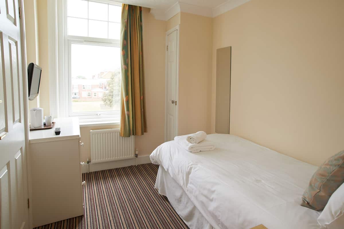 Een hotelkamer met een bed, een nachtkastje met een beker en afstandsbediening, een raam met gordijnen en een radiator.