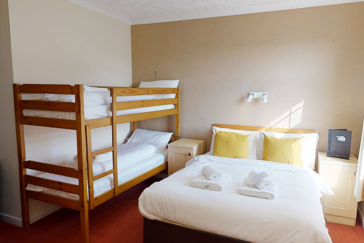 Een hotelkamer met twee houten stapelbedden, een enkel bed met wit beddengoed, en een klein nachtkastje met een lamp.