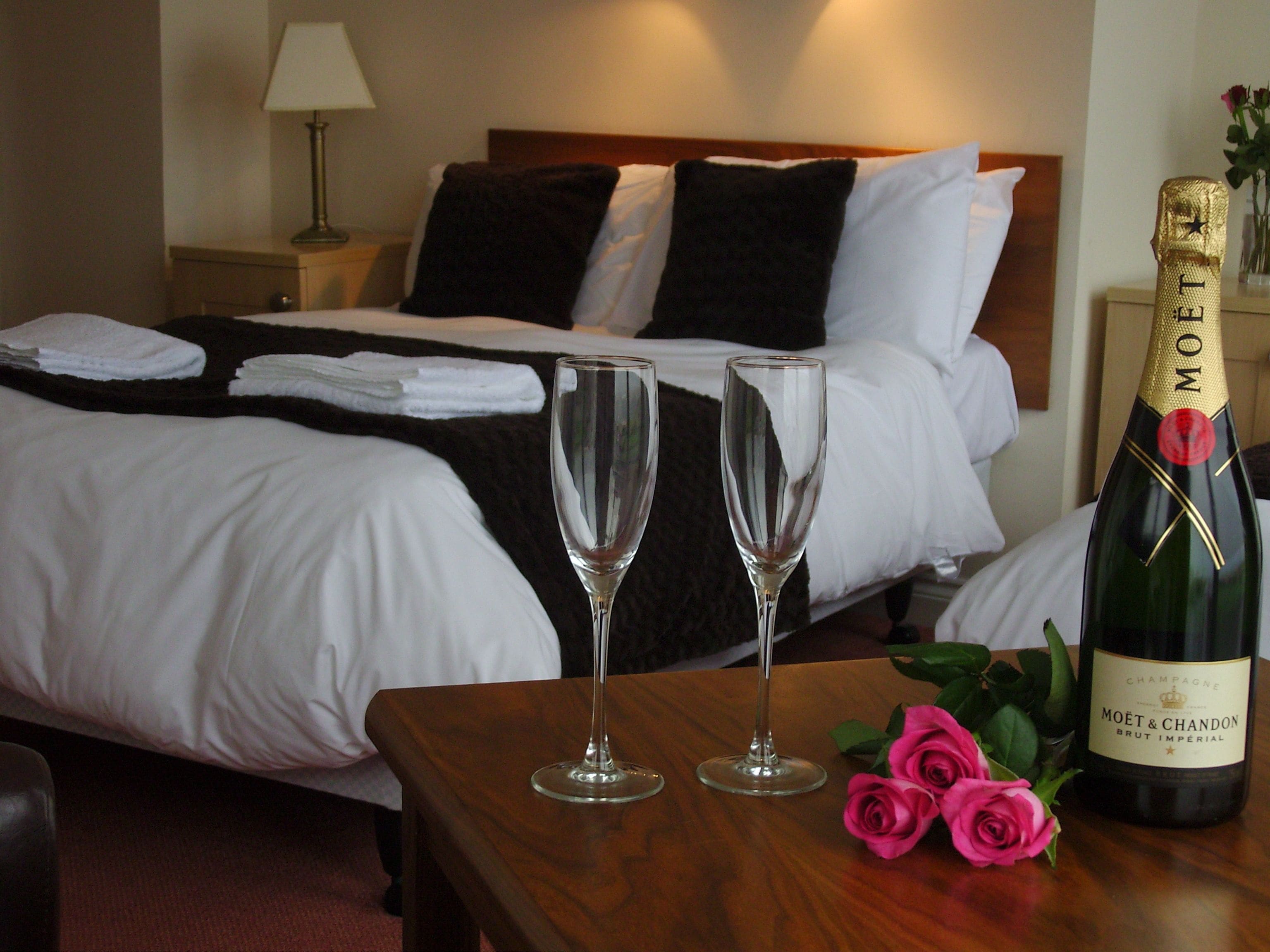 A hotel room with a neatly made bed, two empty wine glasses, a bottle of Moët & Chandon Brut Imperial champagne, and a bouquet of pink roses on a wooden table.