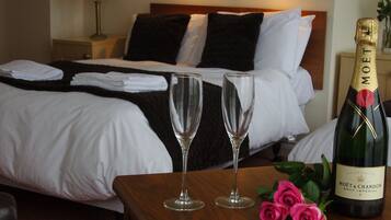A hotel room with a neatly made bed, two empty wine glasses, a bottle of Moët & Chandon Brut Imperial champagne, and a bouquet of pink roses on a wooden table.