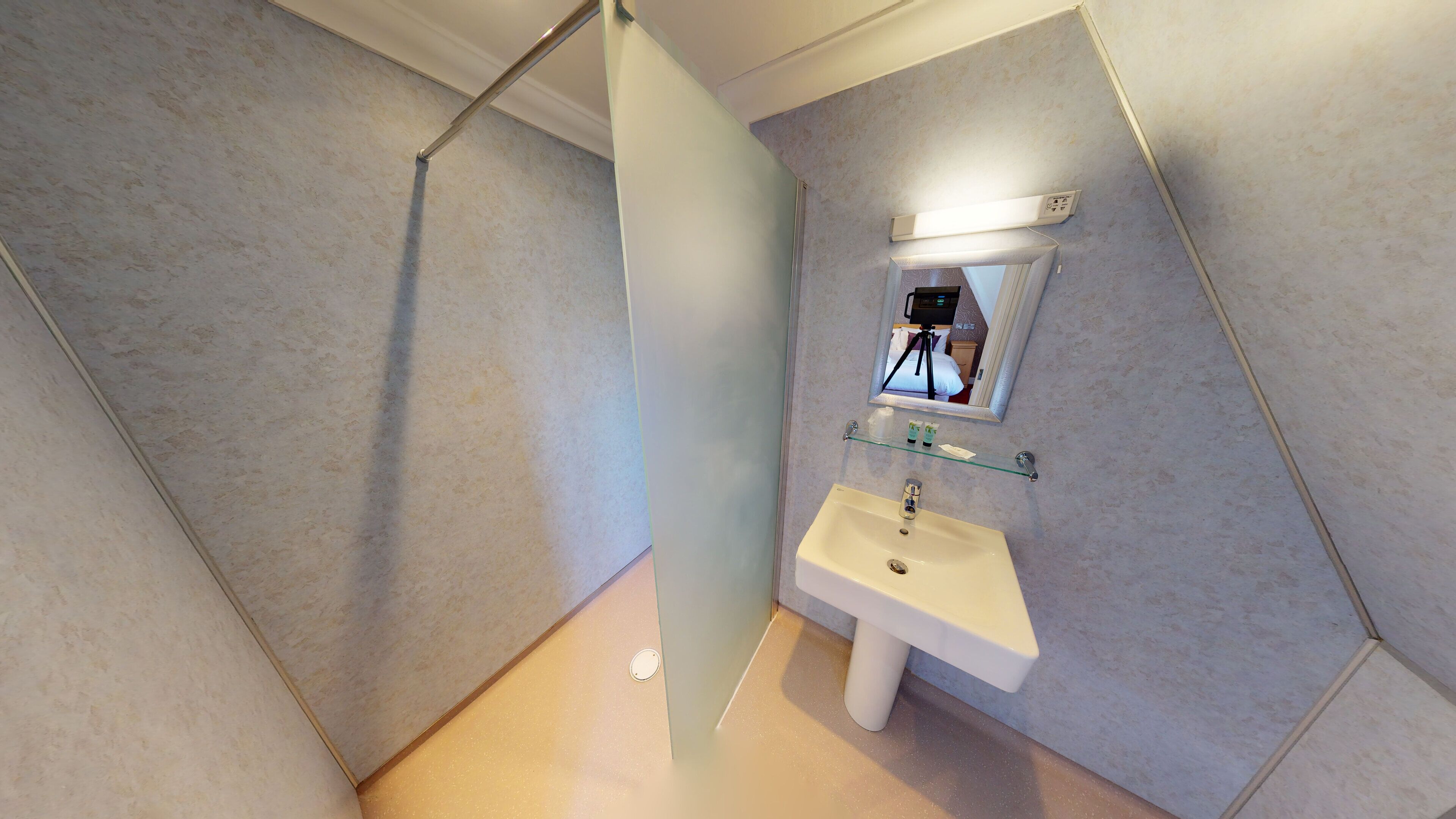 Een moderne badkamer met een witte wastafel, een glazen plank erboven en een spiegel die een bed met een statief weerkaatst.