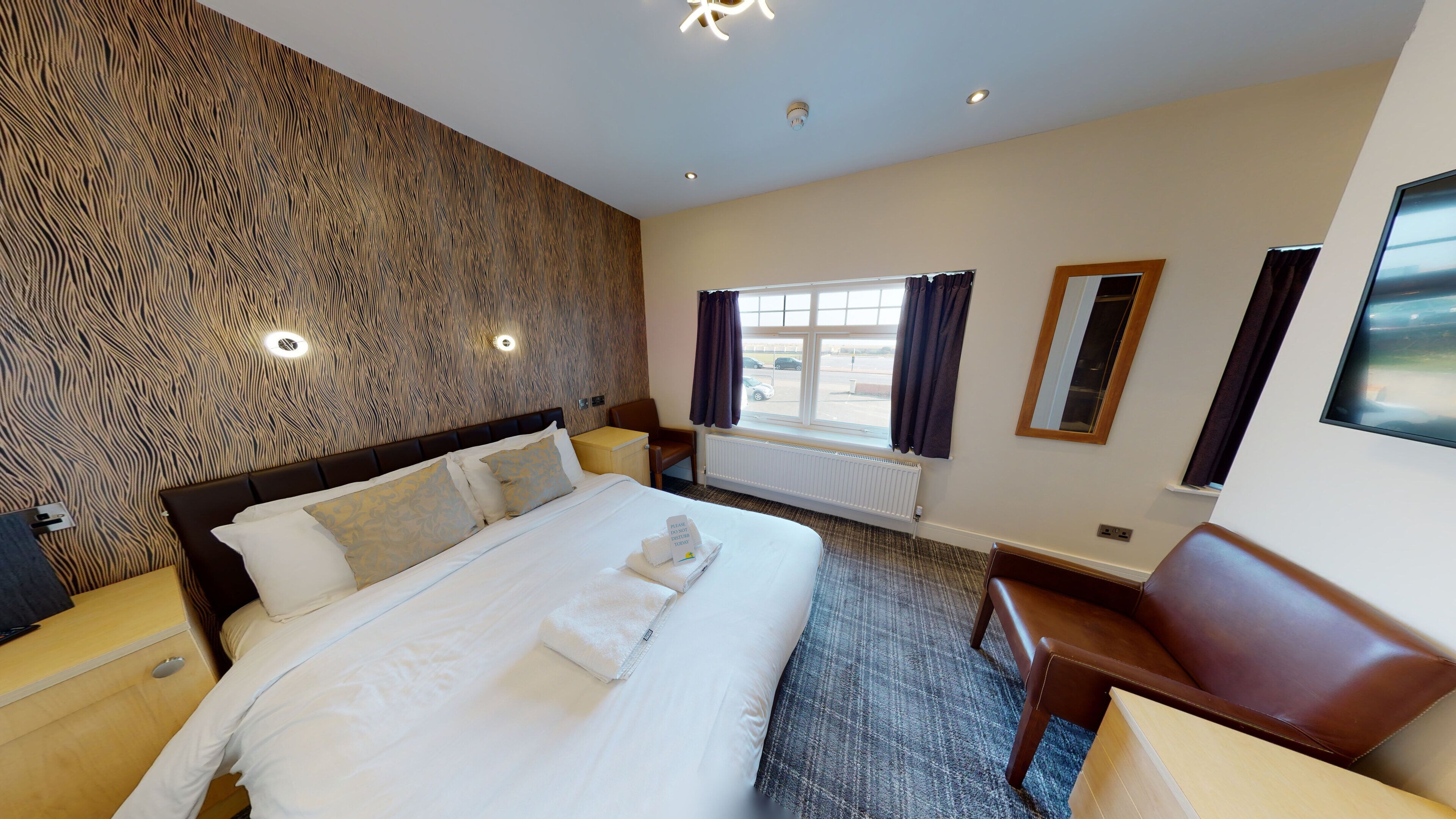 A hotel room with a large bed, a wooden accent wall, a window with curtains, a desk, and a chair.