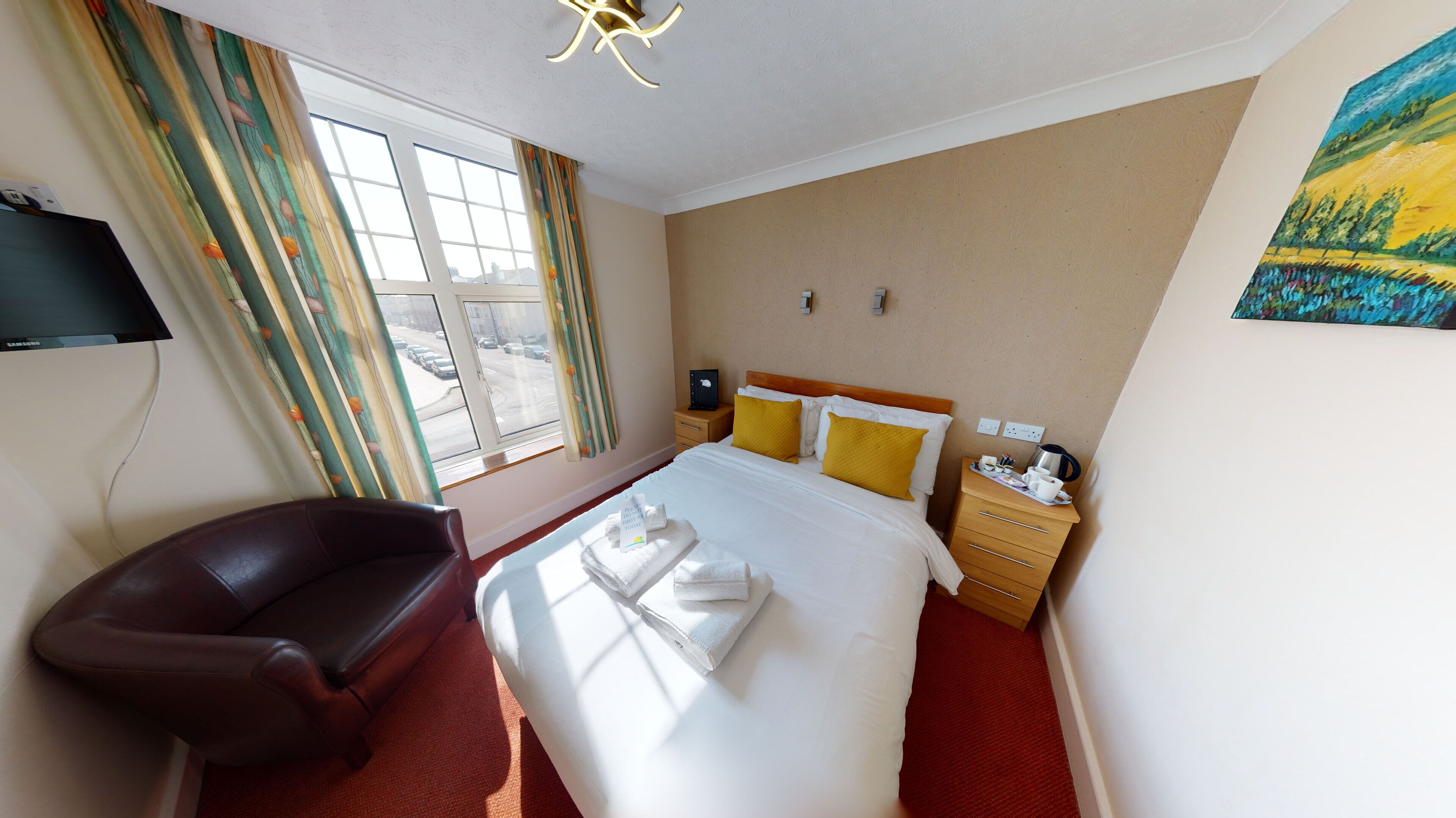Ein Hotelzimmer mit Bett, Sessel, Nachttisch, einem Bild und einem Fenster mit Vorhängen.