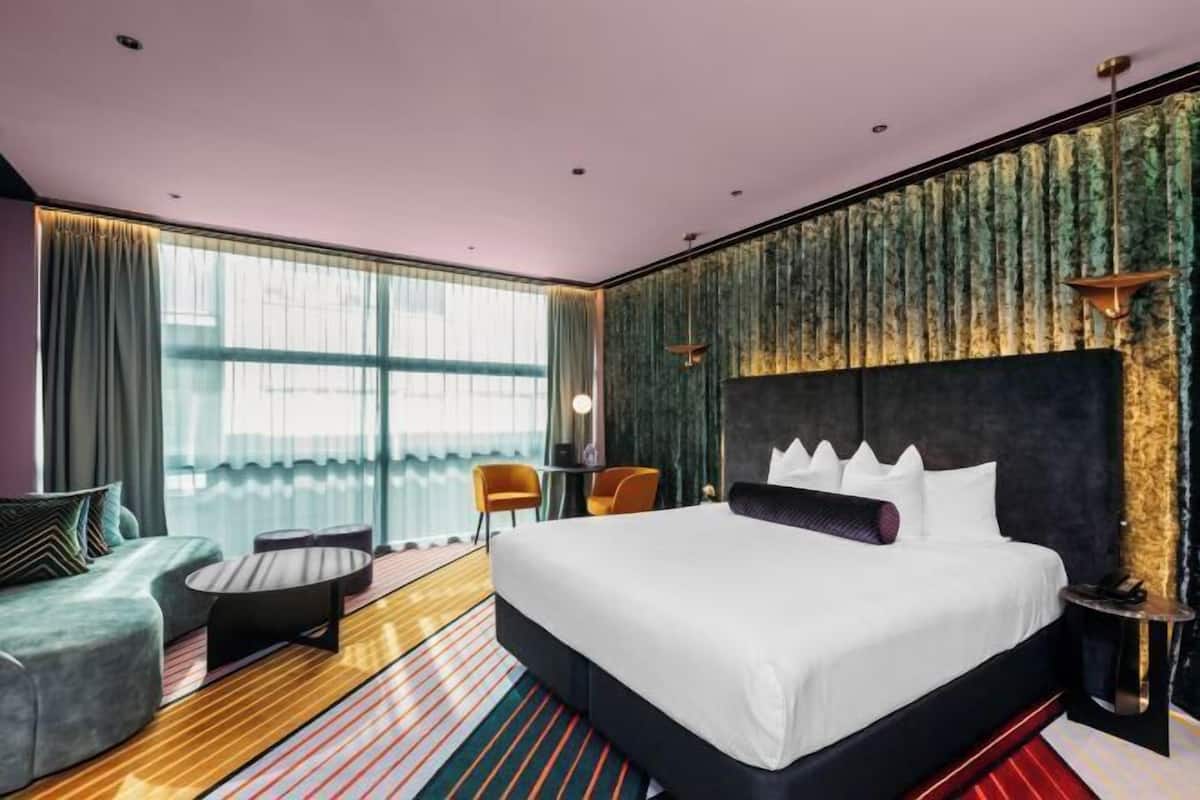 A modern hotel room with a large bed, a sofa, a small table, and a view of the outdoors.