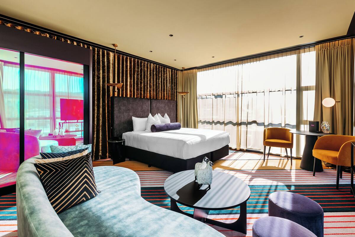 A modern hotel room with a large bed, a sofa, a coffee table, and a view of the city through large windows.