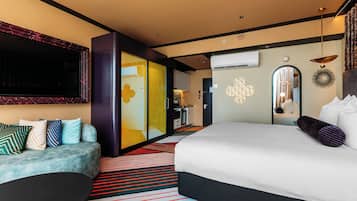 Quarto de hotel moderno com uma cama grande, área de estar com sofá, escrivaninha e um espelho amplo.